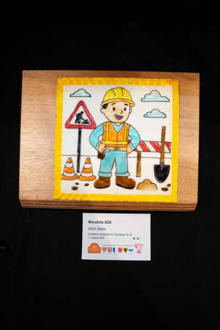 Ein farbenfrohes, essbares Kunstwerk zeigt einen lächelnden Bauarbeiter mit Werkzeugen, Verkehrsschildern und Leitkegeln auf einem Holzsockel. Ein Schild darunter enthält Informationen zum Künstler und zum Wettbewerb.