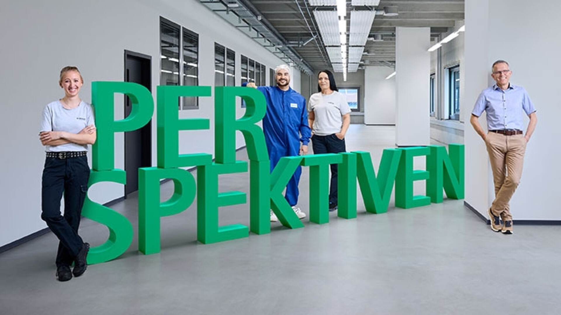 Vier Personen stehen in einem hellen, modernen Flur neben großen grünen 3D-Buchstaben mit dem Wort „PERSPEKTIVEN“. Sie lächeln und posieren lässig und stehen für Vielfalt und Teamwork.