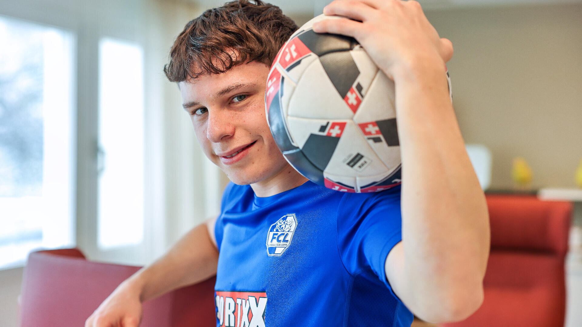 Ein junger Mensch in einem blauen Fußballtrikot hält einen Fußball auf der Schulter und lächelt in die Kamera in einem hell erleuchteten Raum.