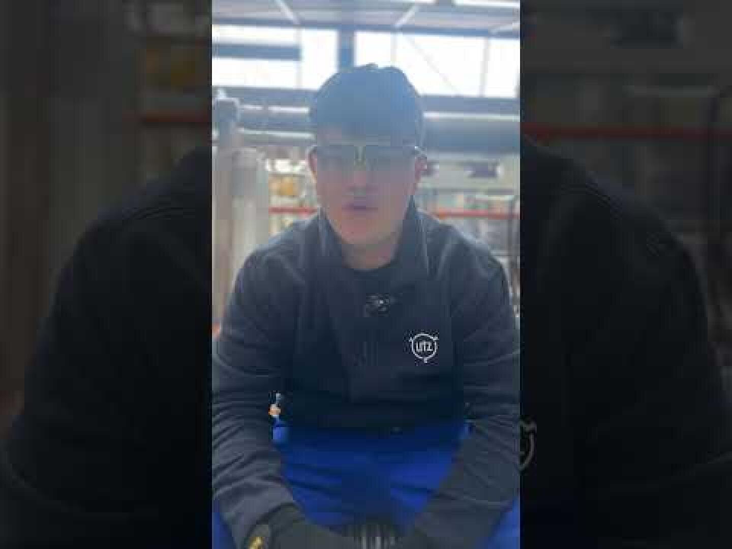 Eine Person, die eine Schutzbrille, Handschuhe und ein dunkles Sweatshirt mit einem Logo trägt, sitzt in einem Innenraum, der wie eine Industrie- oder Werkstatthalle aussieht.