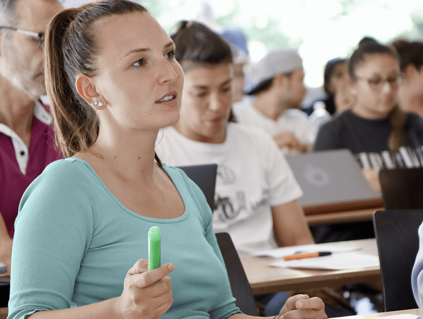 Eine junge Frau, die einen grünen Textmarker in der Hand hält, sitzt aufmerksam in einem Klassenzimmer inmitten anderer Schüler, von denen einige Notizen machen oder Laptops benutzen.
