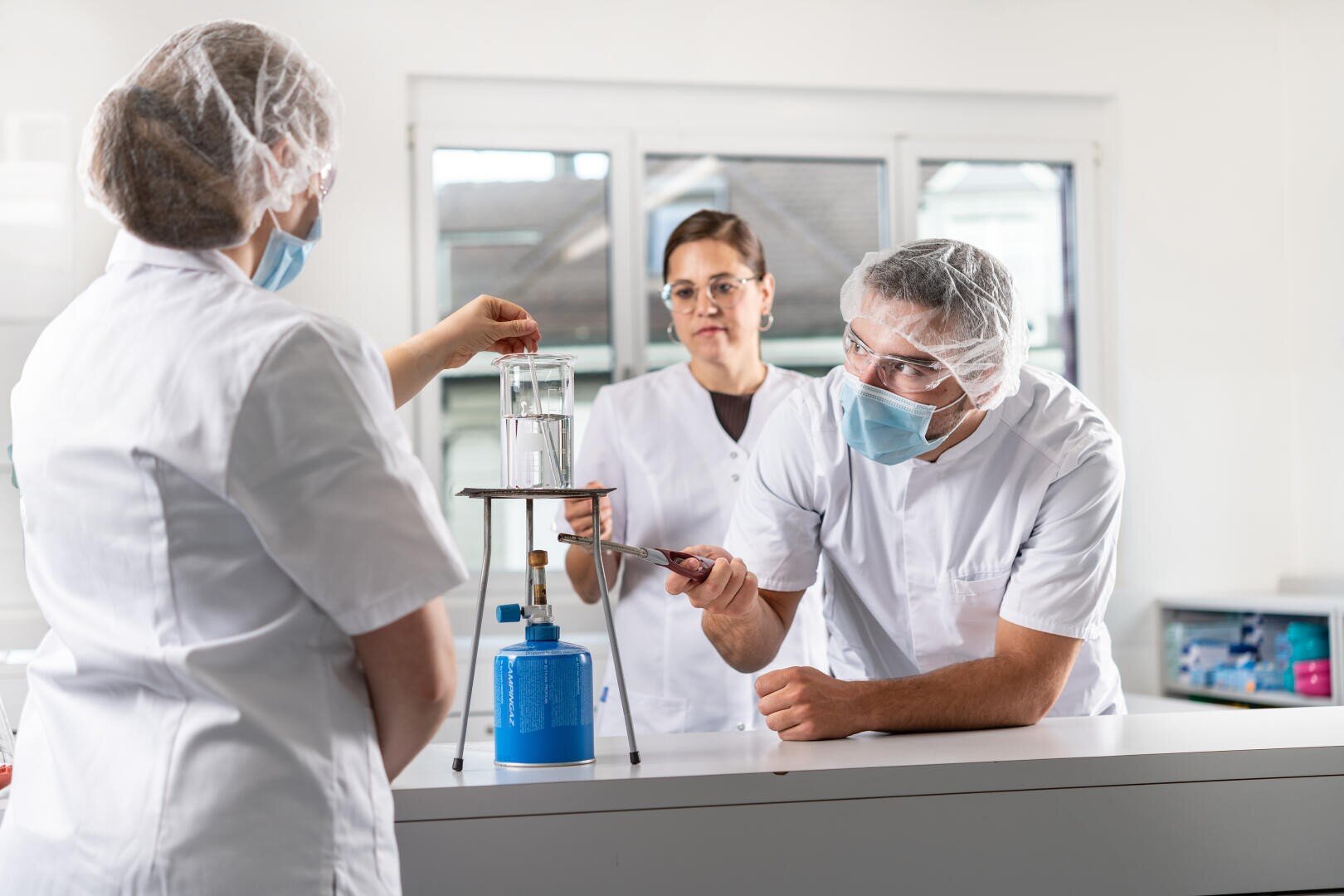 Drei Personen in Laborkitteln, Haarnetzen und Gesichtsmasken führen in einem hellen Labor ein Experiment mit Glasgeräten und einem Bunsenbrenner auf einer Arbeitsplatte durch.