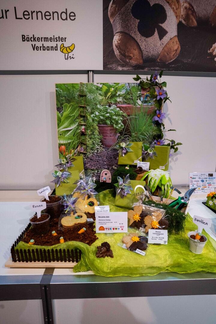 Eine farbenfrohe Bäckerei-Auslage zeigt eine Gartenszene mit Blumen, kleinen Schildern, Pflanzen und Backwaren. Im Hintergrund ist ein echter Garten zu sehen, und der Tisch ist mit einem grünen Tischtuch und Miniatur-Gartenaccessoires dekoriert.