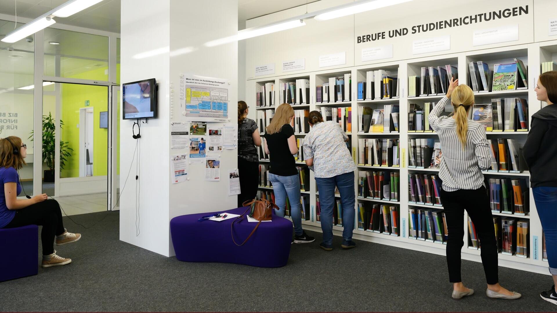 In einer modernen Bibliothek oder einem Karrierezentrum schauen sich mehrere Personen Bücher und Broschüren in Regalen mit der Aufschrift „Berufe und Studienrichtungen“ an. Eine Frau sitzt in der Nähe eines Fernsehbildschirms, während andere die Materialien durchblättern.