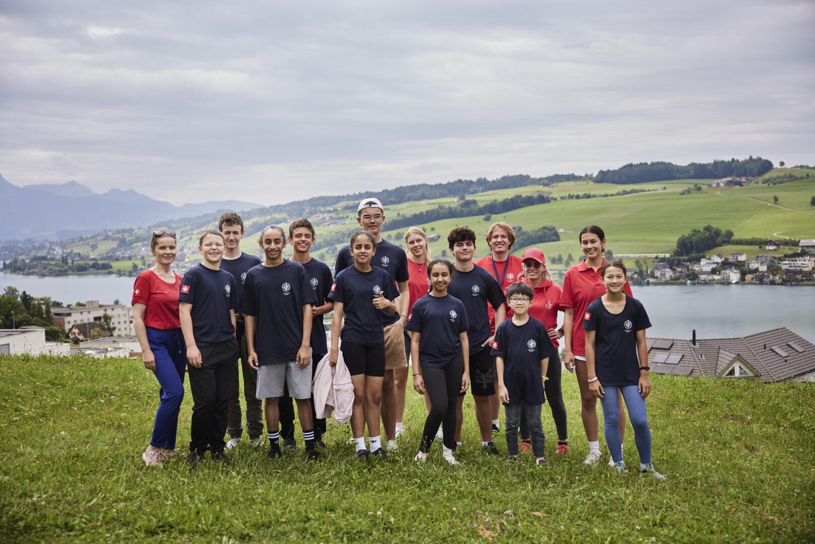 Eine Gruppe Jugendlicher und Erwachsener posiert zusammen auf einem grasbewachsenen Hügel mit einem See und Häusern im Hintergrund. Sie tragen passende marineblaue und rote Hemden. Die Szene ist im Freien mit grünen Hügeln und Bergen unter einem bewölkten Himmel.