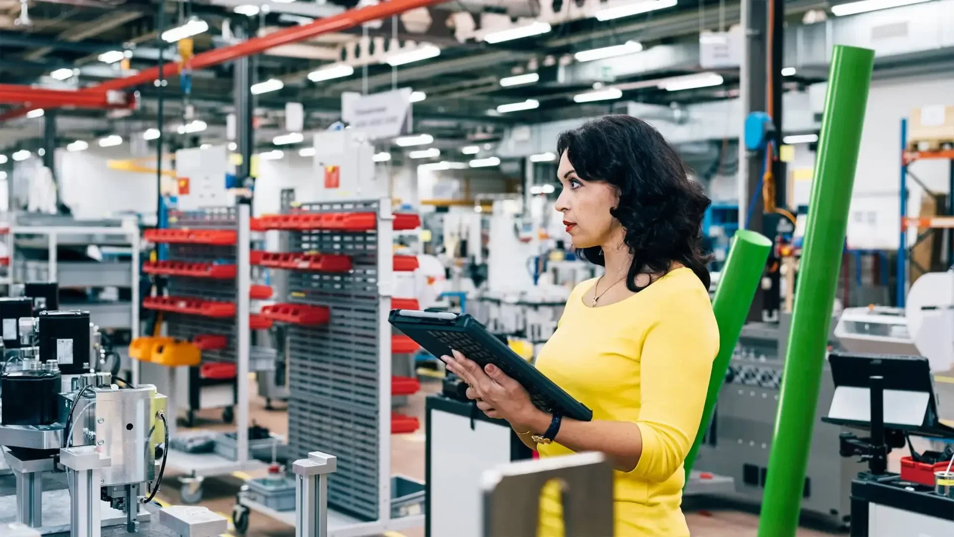 Eine Frau in einem gelben Hemd hält ein digitales Tablet in der Hand, während sie in einer hell erleuchteten, gut organisierten Fabrik oder Produktionsstätte mit Regalen und Werkzeugen im Hintergrund die Ausrüstung überprüft.