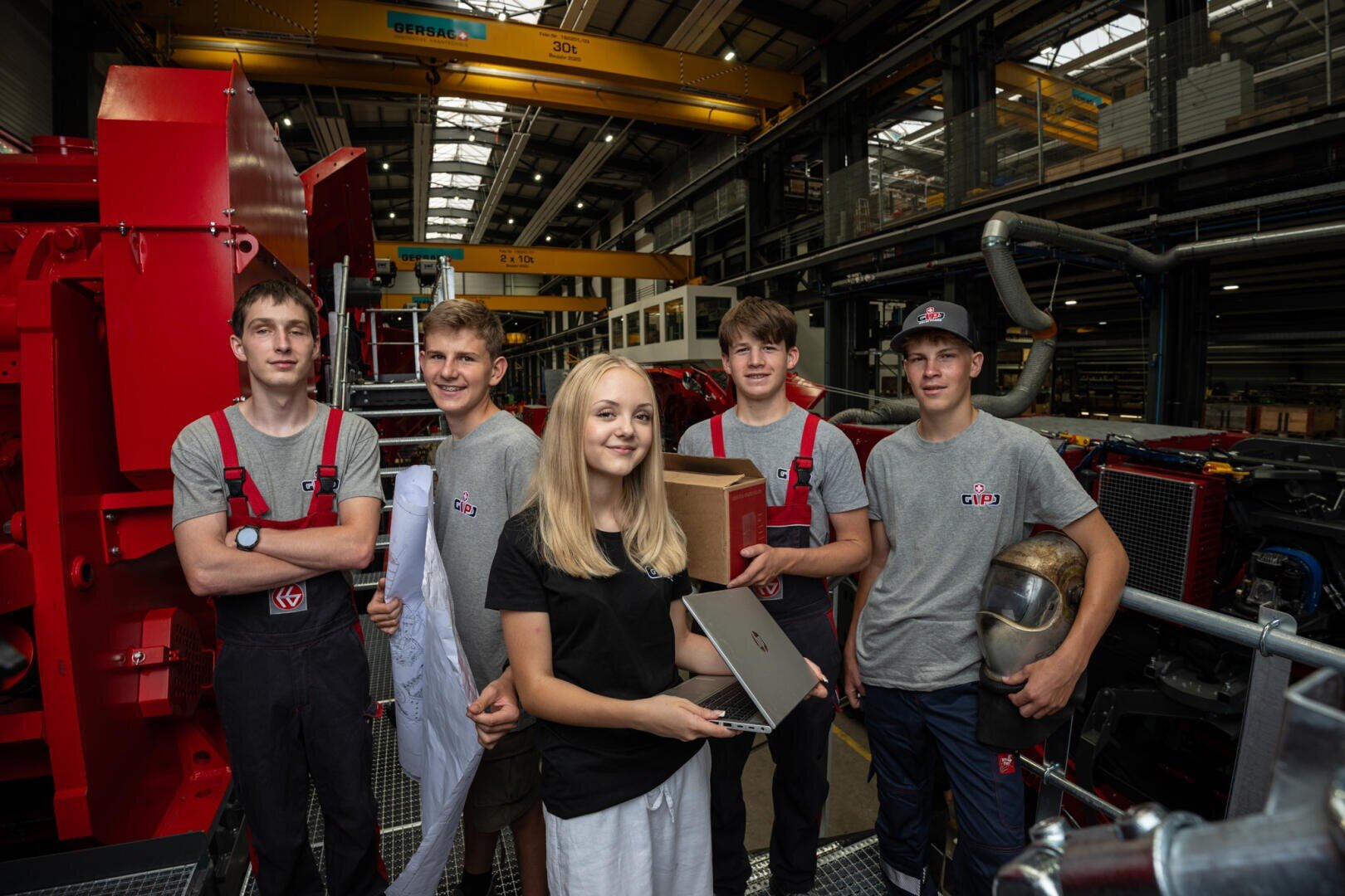 Fünf junge Menschen stehen in einer Fabrikhalle; einige tragen graue Hemden und halten Geräte wie einen Laptop und eine Kiste in den Händen, im Hintergrund sind Industriemaschinen und gelbe Kräne zu sehen.