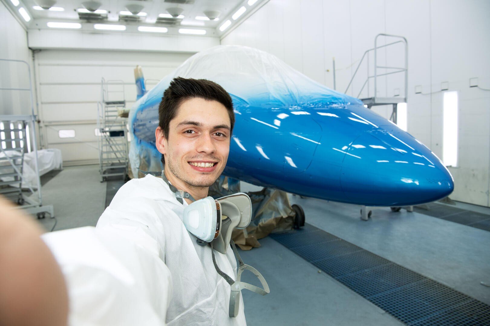 Ein Mann in Schutzkleidung macht ein Selfie vor einem blau-weißen Flugzeug in einem hell erleuchteten Hangar oder einer Werkstatt.