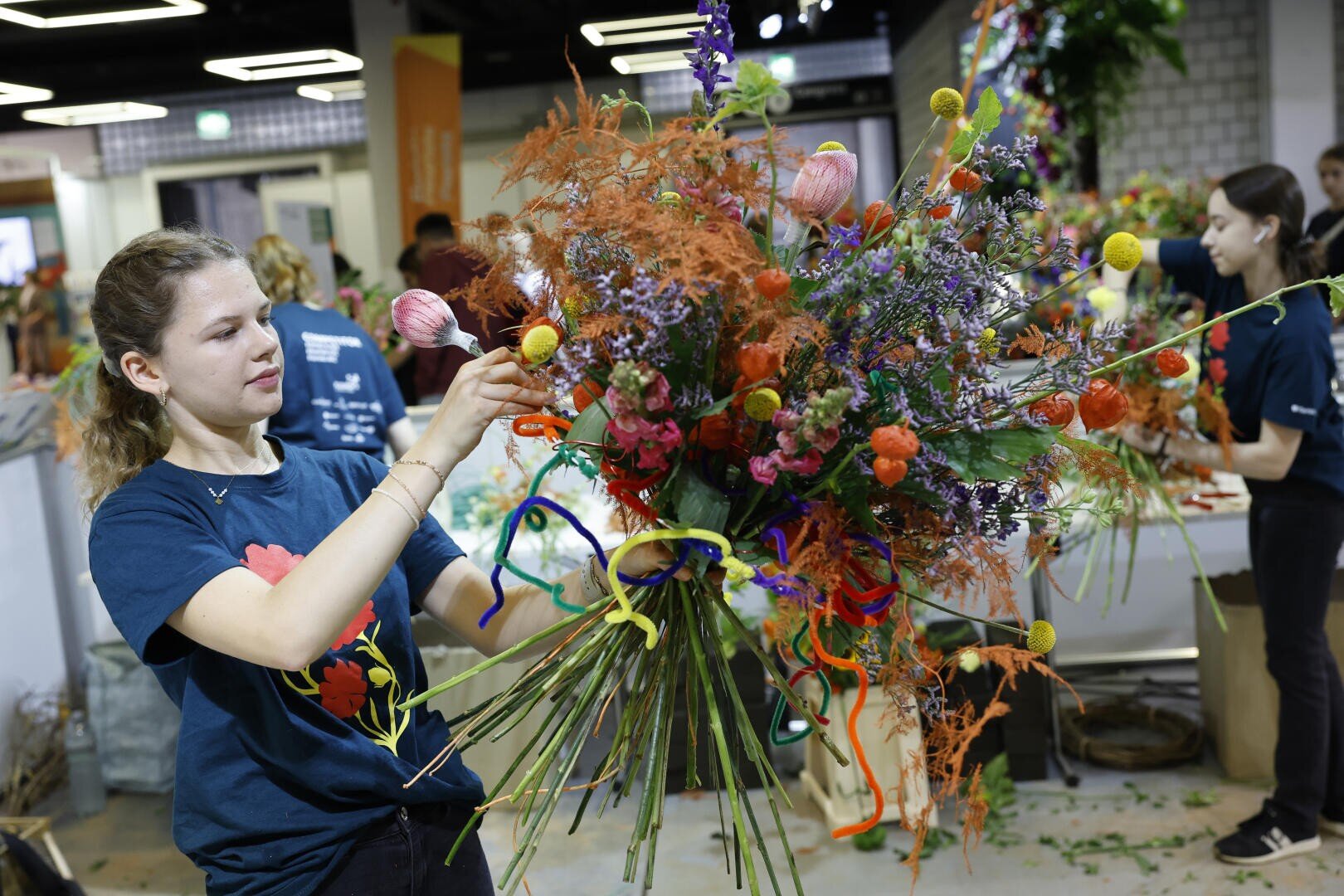 Eine junge Frau arrangiert einen großen, farbenfrohen Strauß mit verschiedenen Blumen und Grünpflanzen bei einer Indoor-Veranstaltung, während andere im Hintergrund an Blumenarrangements arbeiten.