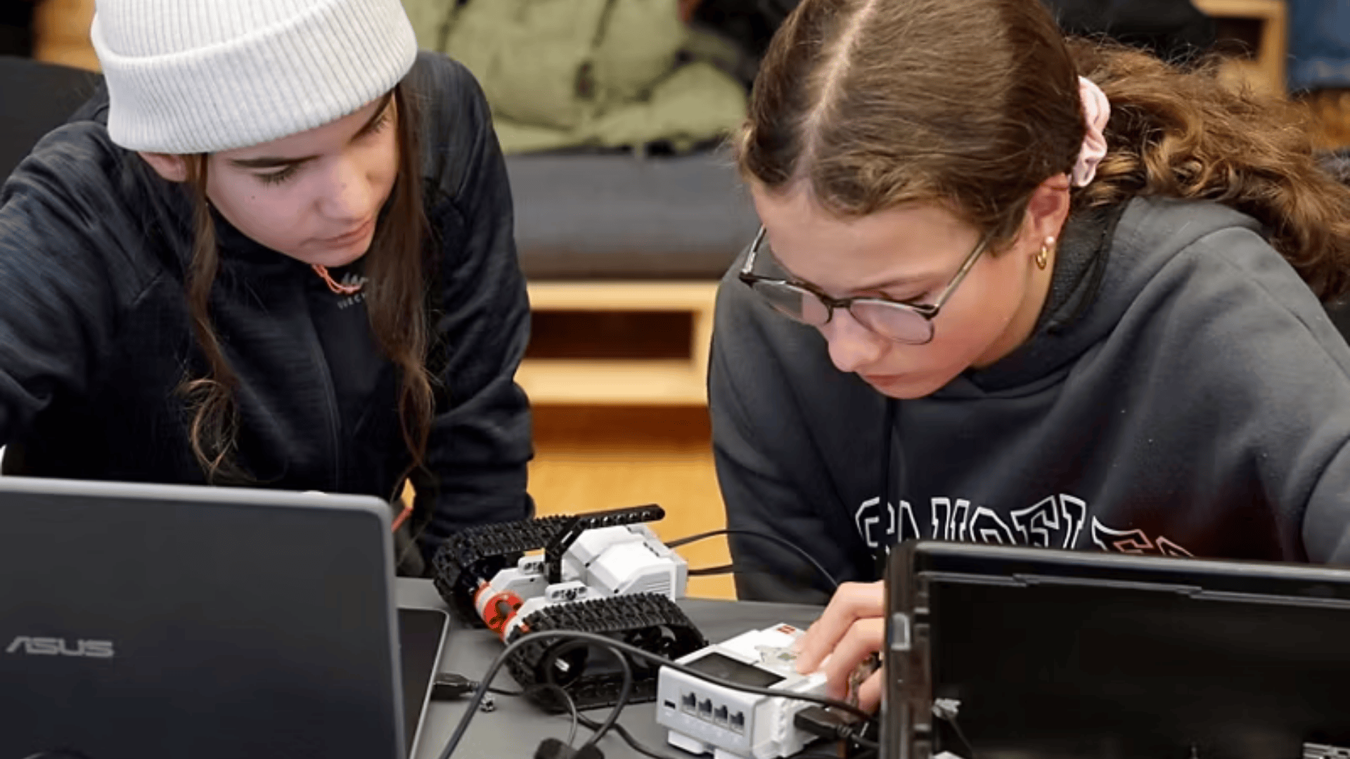 Zwei junge Leute arbeiten konzentriert an einem Tisch mit Laptops, elektronischen Bauteilen und einem kleinen Roboter und konzentrieren sich darauf, das Gerät miteinander zu verbinden oder zu programmieren.