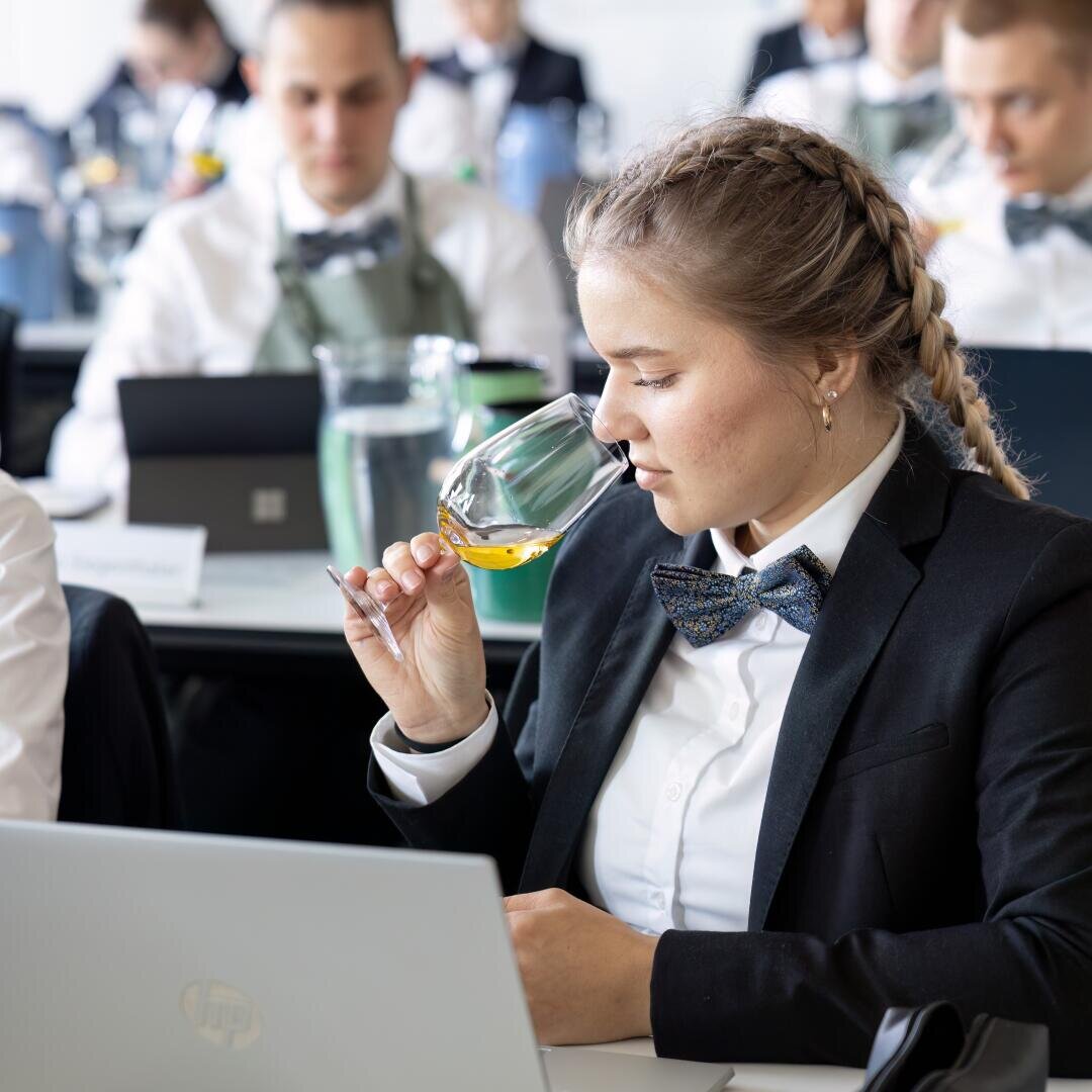 Eine Frau in Anzug und Fliege sitzt an einem Schreibtisch und riecht an einem Glas mit gelber Flüssigkeit. Sie befindet sich in einem Klassenzimmer mit anderen förmlich gekleideten Personen, von denen einige mit Laptops arbeiten. Die Umgebung wirkt professionell und konzentriert.