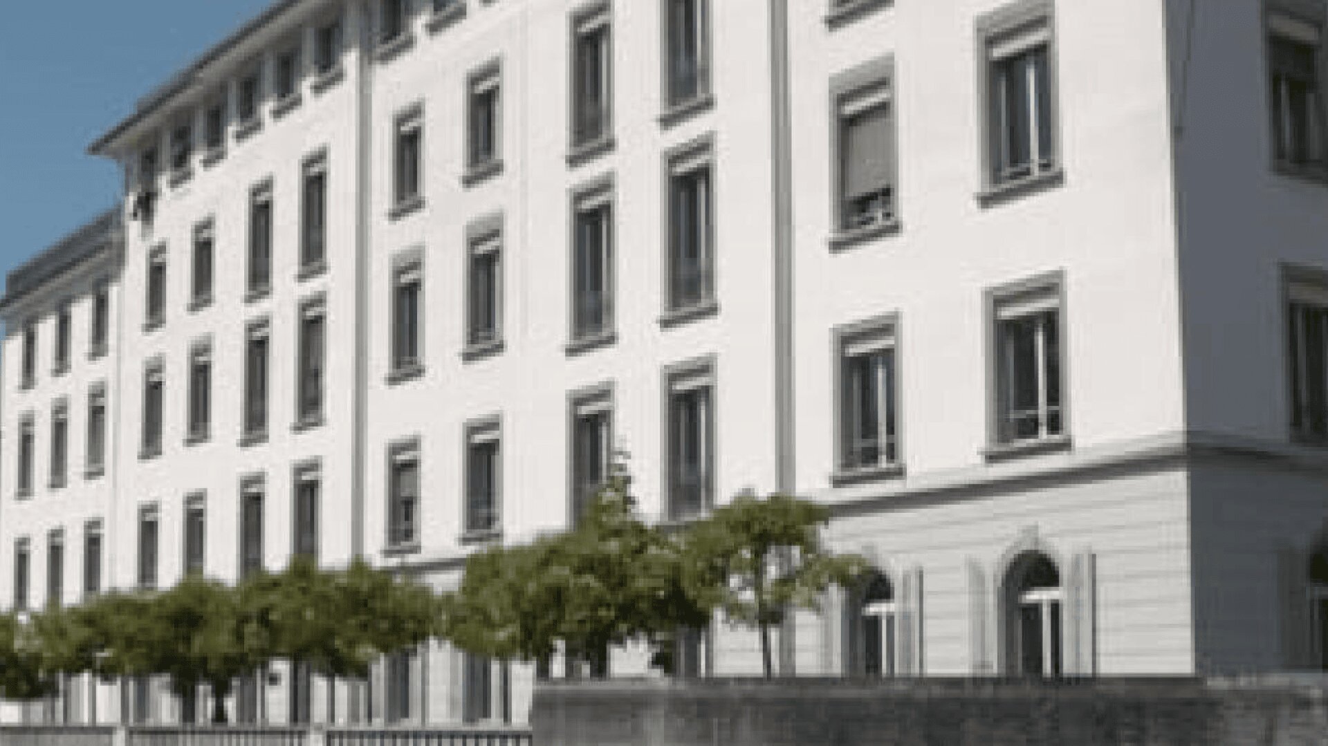 Ein weißes, mehrstöckiges Gebäude mit Reihen von Fenstern und einem grauen Sockel. Kleine Bäume säumen den Gehweg davor, und eine Steinmauer verläuft entlang der unteren Kante des Gebäudes. Der Himmel ist klar und blau.