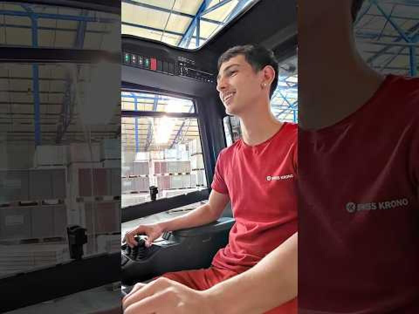 Eine Person in einem roten Hemd bedient Maschinen in einem Lagerhaus, in dessen Hintergrund Materialstapel und eine blau gerahmte Decke zu sehen sind.