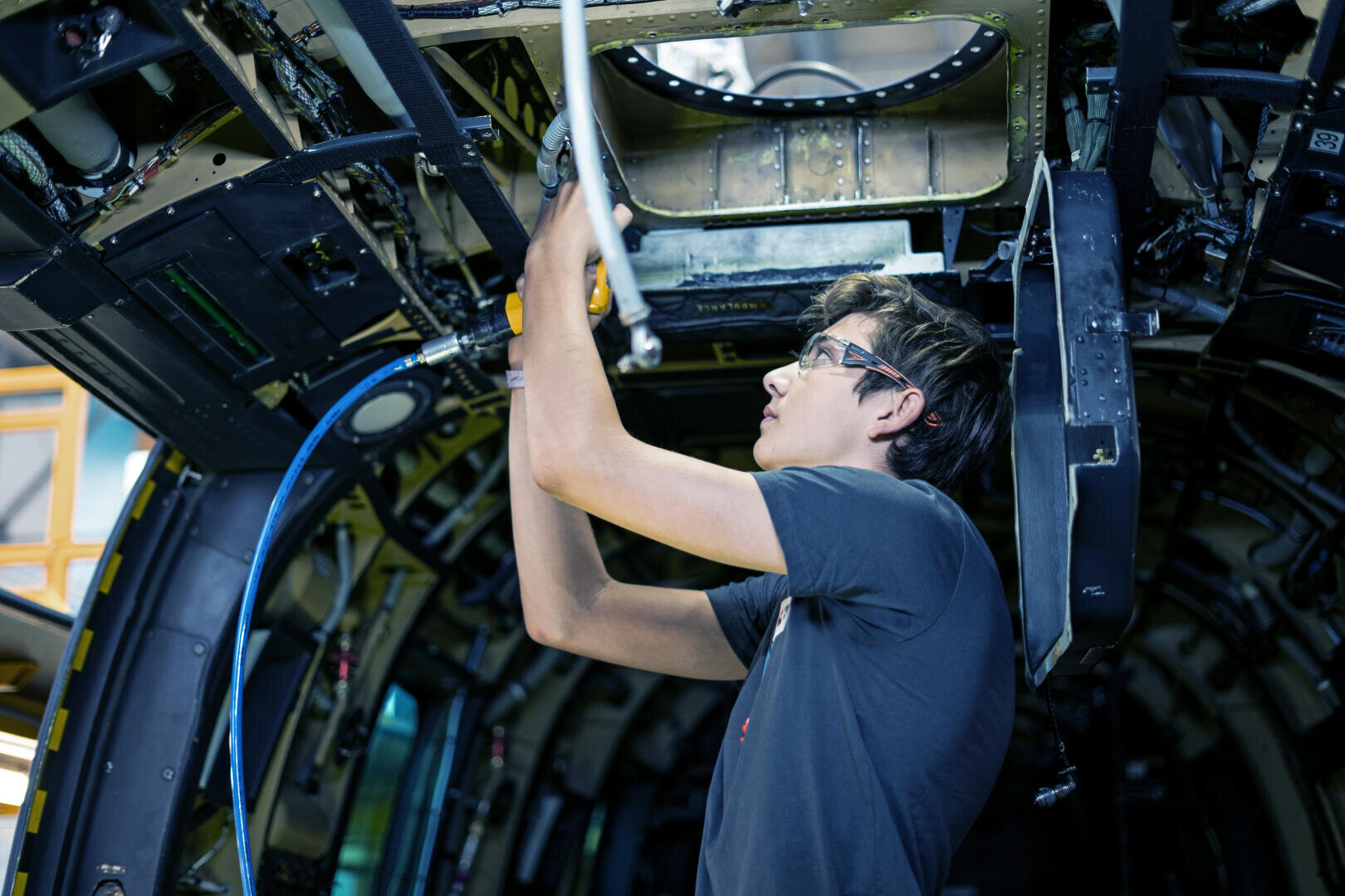 Ein junger Mann mit Schutzbrille arbeitet mit Werkzeugen an der Verdrahtung und den Verkleidungen im Rumpf eines Flugzeugs, umgeben von freiliegenden Bauteilen und komplizierten Maschinen.