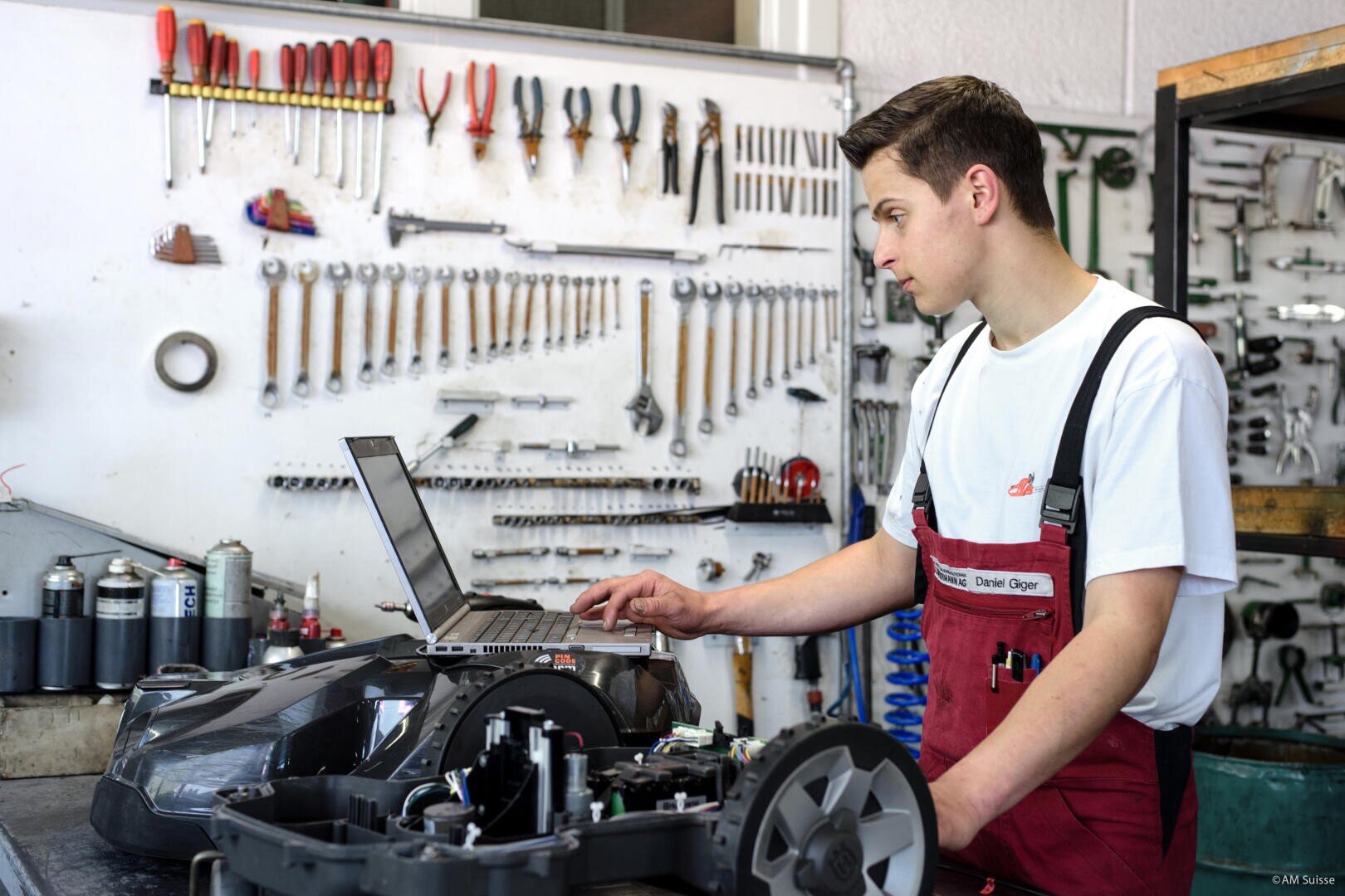 Ein junger Mechaniker im Overall arbeitet an einem Laptop in einer Werkstatt, umgeben von Werkzeugen an der Wand und verschiedenen Teilen auf der Werkbank. Er scheint eine Maschine zu diagnostizieren oder zu reparieren.