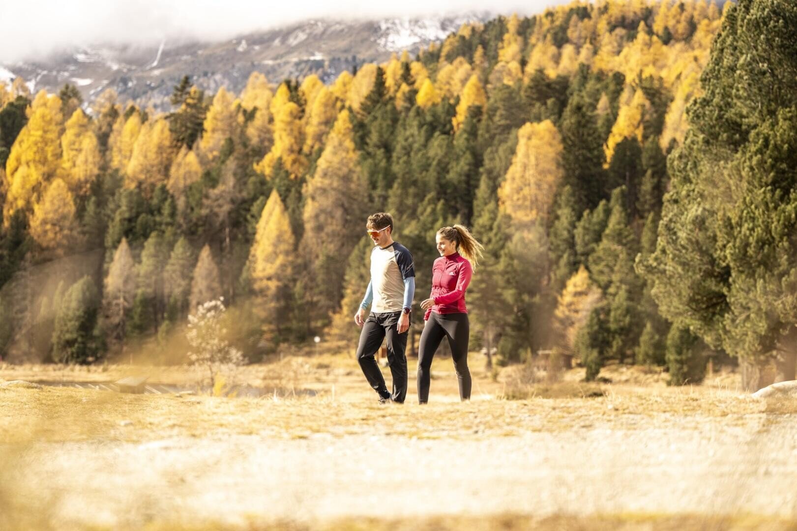 Zwei Tourismusstudierende der HF Tourismus & Management in Samedan spazieren gemeinsam über eine Wiese, im Hintergrund sind herbstlich gefärbte Bäume und Berge zu sehen, und genießen einen sonnigen Tag im Freien.