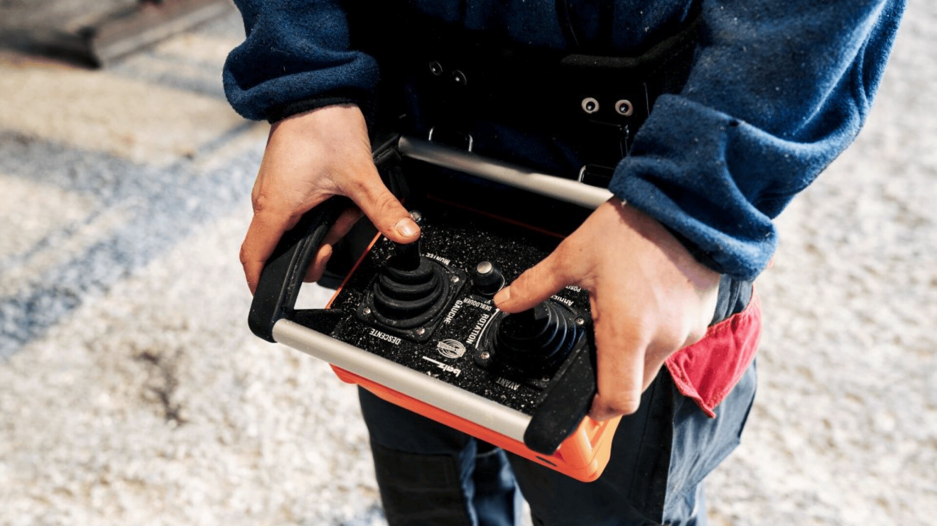 Eine Person, die ein blaues, langärmeliges Hemd trägt, bedient eine tragbare Fernsteuerung mit zwei Joysticks und steht dabei auf einer hellen, strukturierten Fläche.