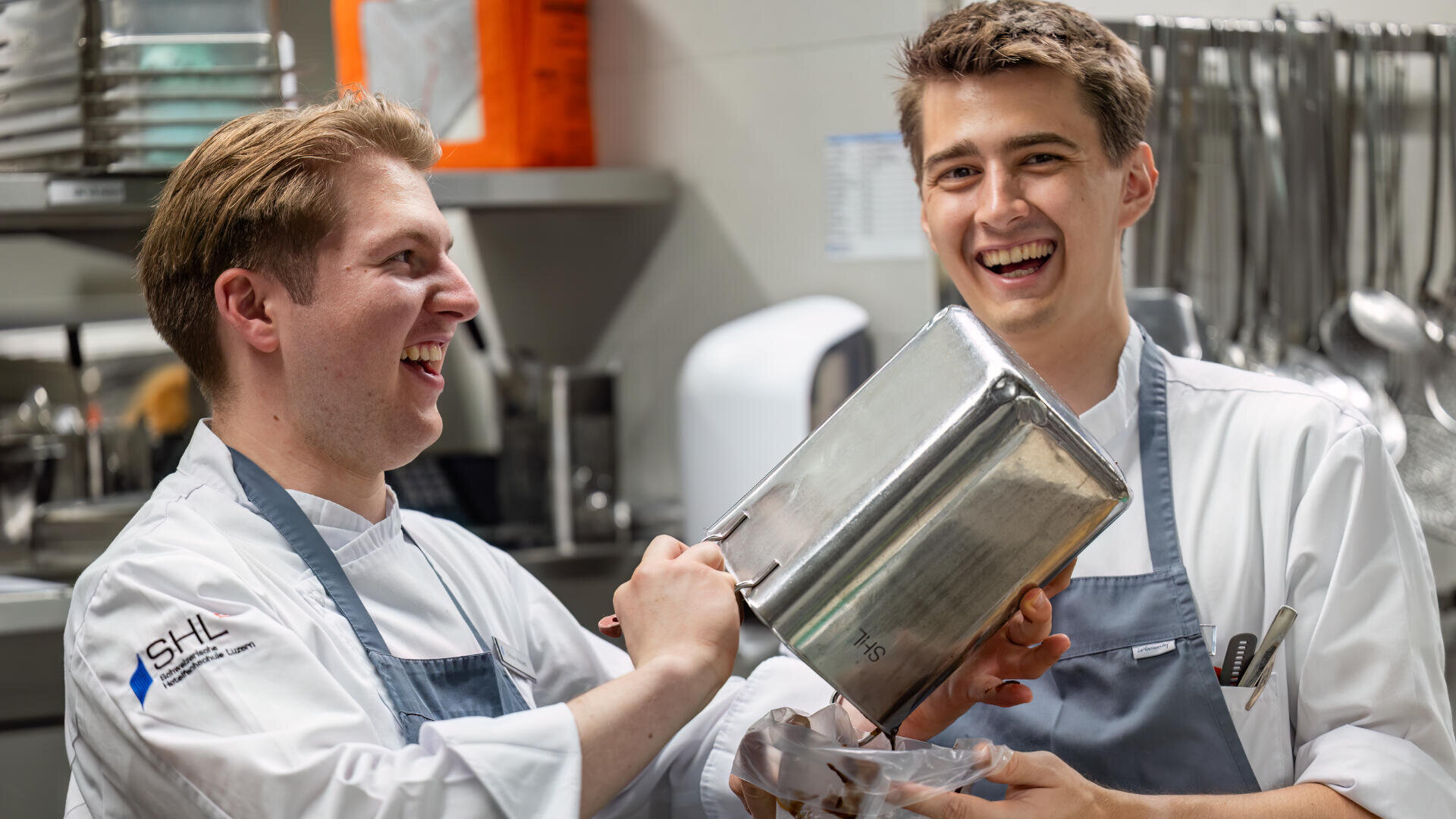Zwei junge männliche Köche in weißen Uniformen und Schürzen lächeln und lachen in einer professionellen Küche, während sie Essen aus einem großen Metallbehälter einfüllen. Im Hintergrund sind Kochutensilien zu sehen.