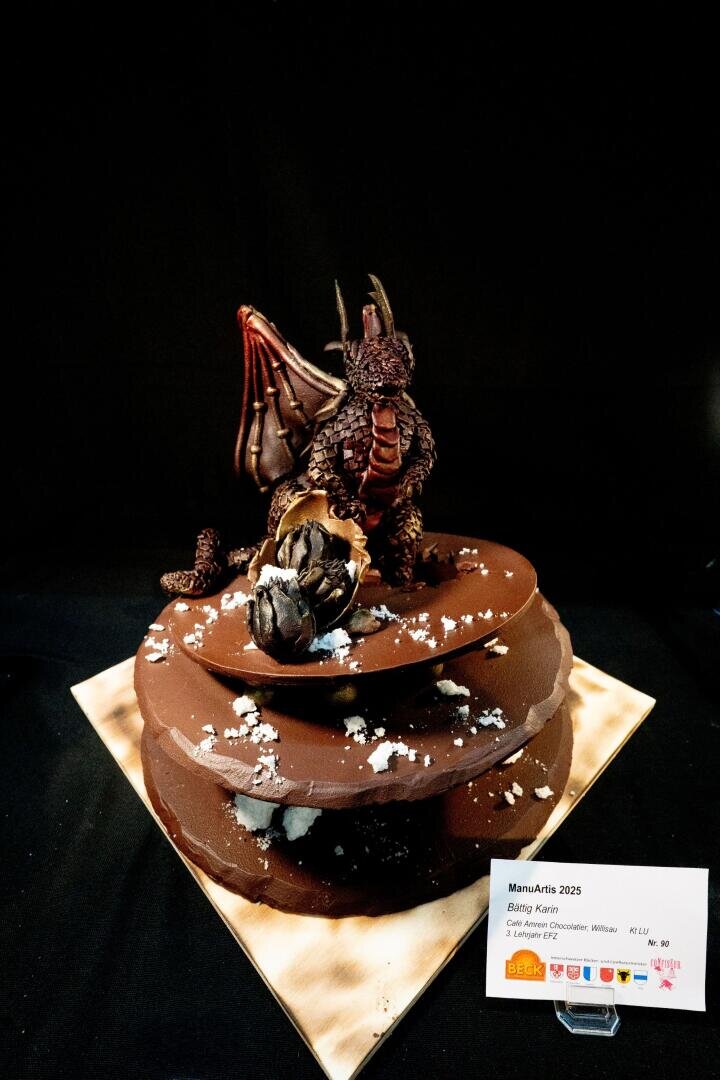 Ein Schokoladenkuchen in Form von aufeinander gestapelten Steinen ist oben mit einem modellierten Drachen mit Flügeln und strukturierten Schuppen verziert. Neben dem Drachen liegt ein aufgebrochenes Schokoladenei. Der Kuchen steht auf einem Holzbrett vor einem schwarzen Hintergrund.
