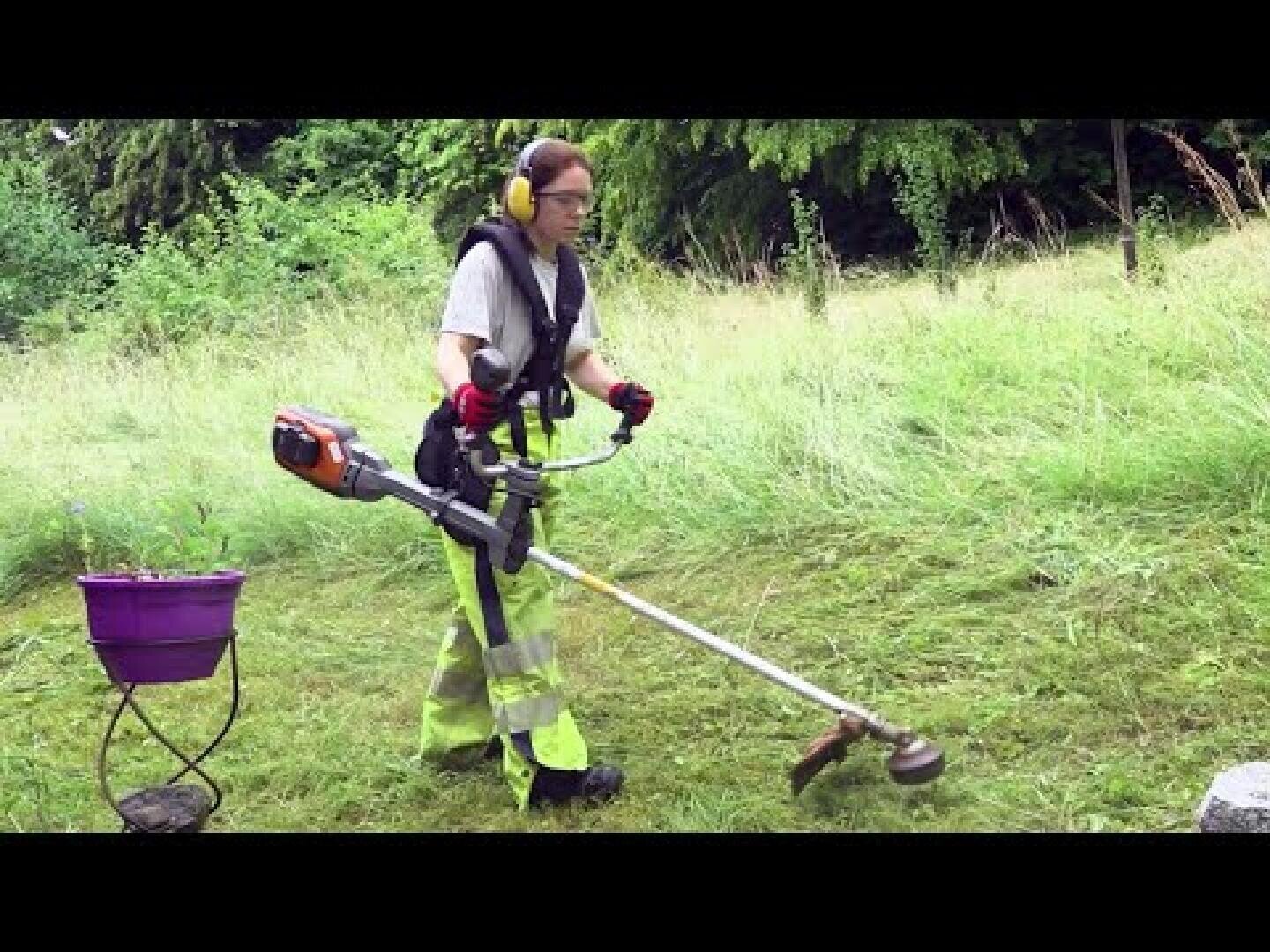 Eine Person, die Schutzkleidung trägt, mäht mit einem Rasentrimmer das Gras in einem Garten. In der Nähe steht ein violetter Eimer auf einem Ständer. Im Hintergrund sind Bäume und hohes Gras zu sehen.