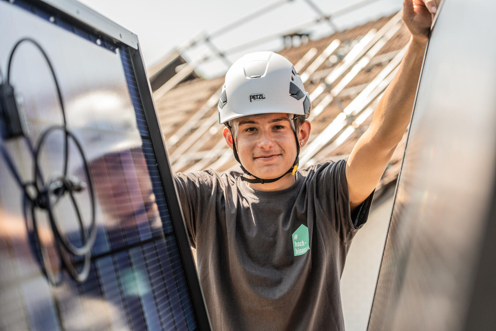 Eine Person, die einen Schutzhelm und ein graues Hemd trägt, lächelt, während sie an einem sonnigen Tag im Freien Solarmodule installiert oder inspiziert.