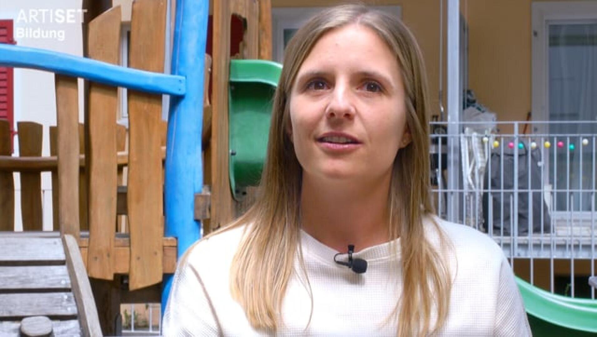 Eine Frau mit langen blonden Haaren und einem weißen Hemd sitzt draußen in der Nähe von Spielgeräten und trägt ein Mikrofon. Im Hintergrund sind ein umzäunter Bereich und bunte Spielgeräte zu sehen.