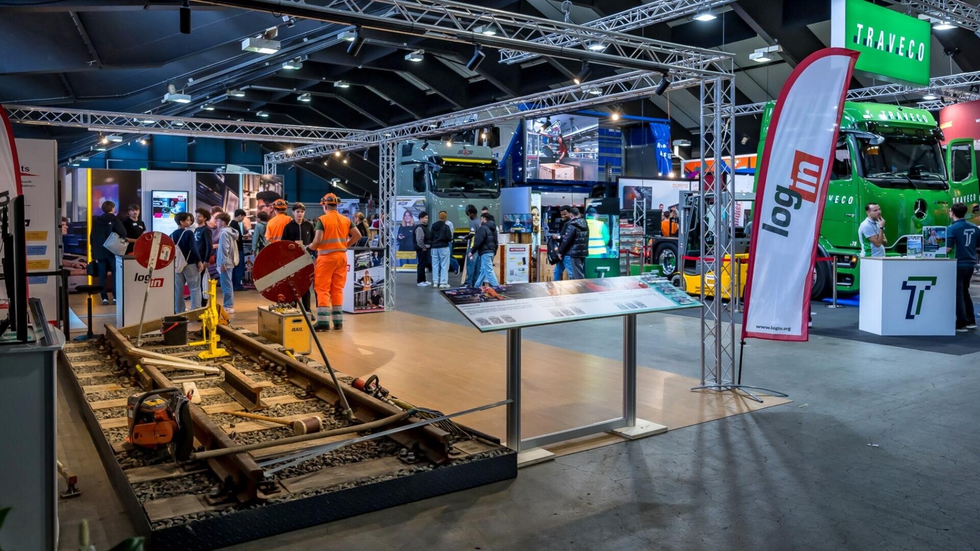 Auf einer überdachten Messe sind Eisenbahnausrüstungen, ein Gleisabschnitt, Informationstafeln, mehrere Stände, Lastwagen und Personengruppen zu sehen, darunter Arbeiter in orangefarbenen Sicherheitsuniformen.