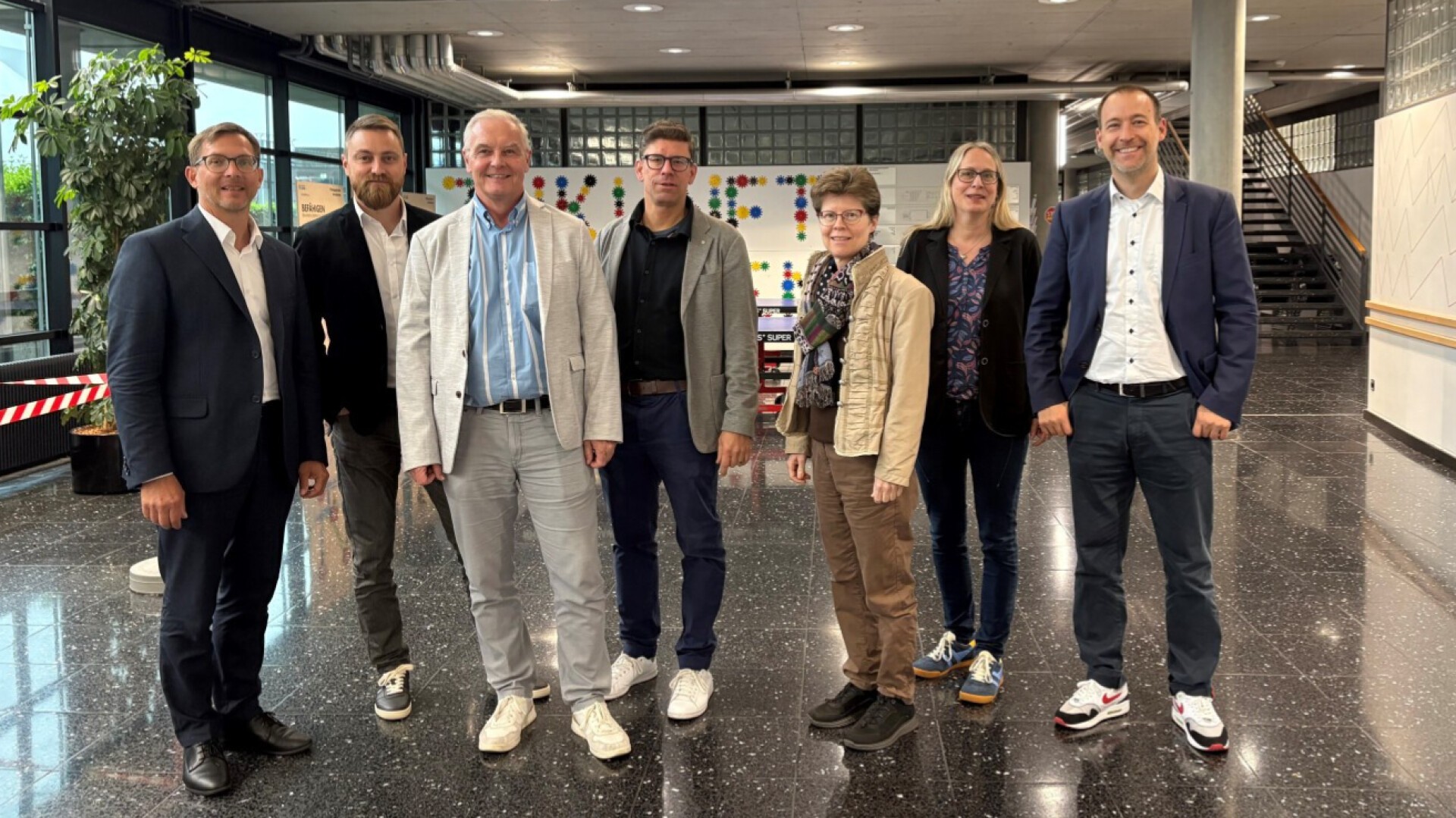 Sieben Personen stehen in einem modernen, geräumigen Gebäude mit glänzend schwarzen Böden und einer Treppe im Hintergrund, posieren und lächeln in die Kamera.