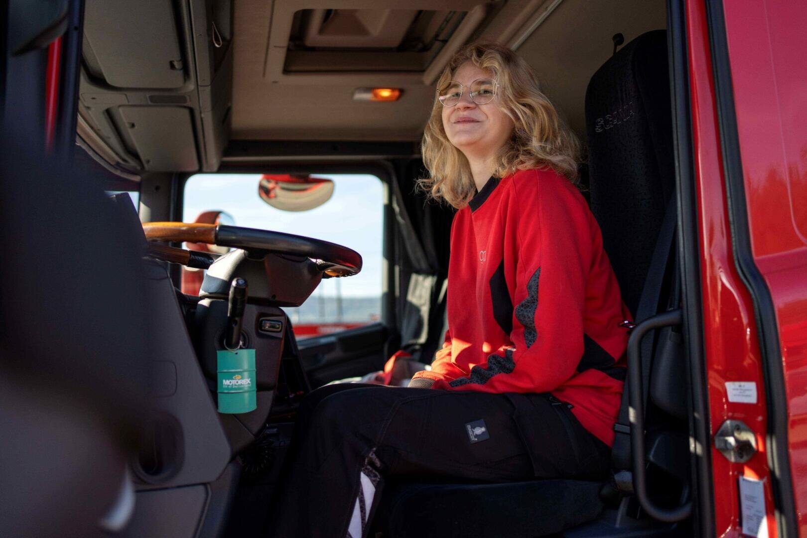 Eine Person mit blonden Haaren, Brille und einem roten Pullover sitzt auf dem Fahrersitz eines roten Lastwagens und lächelt.
