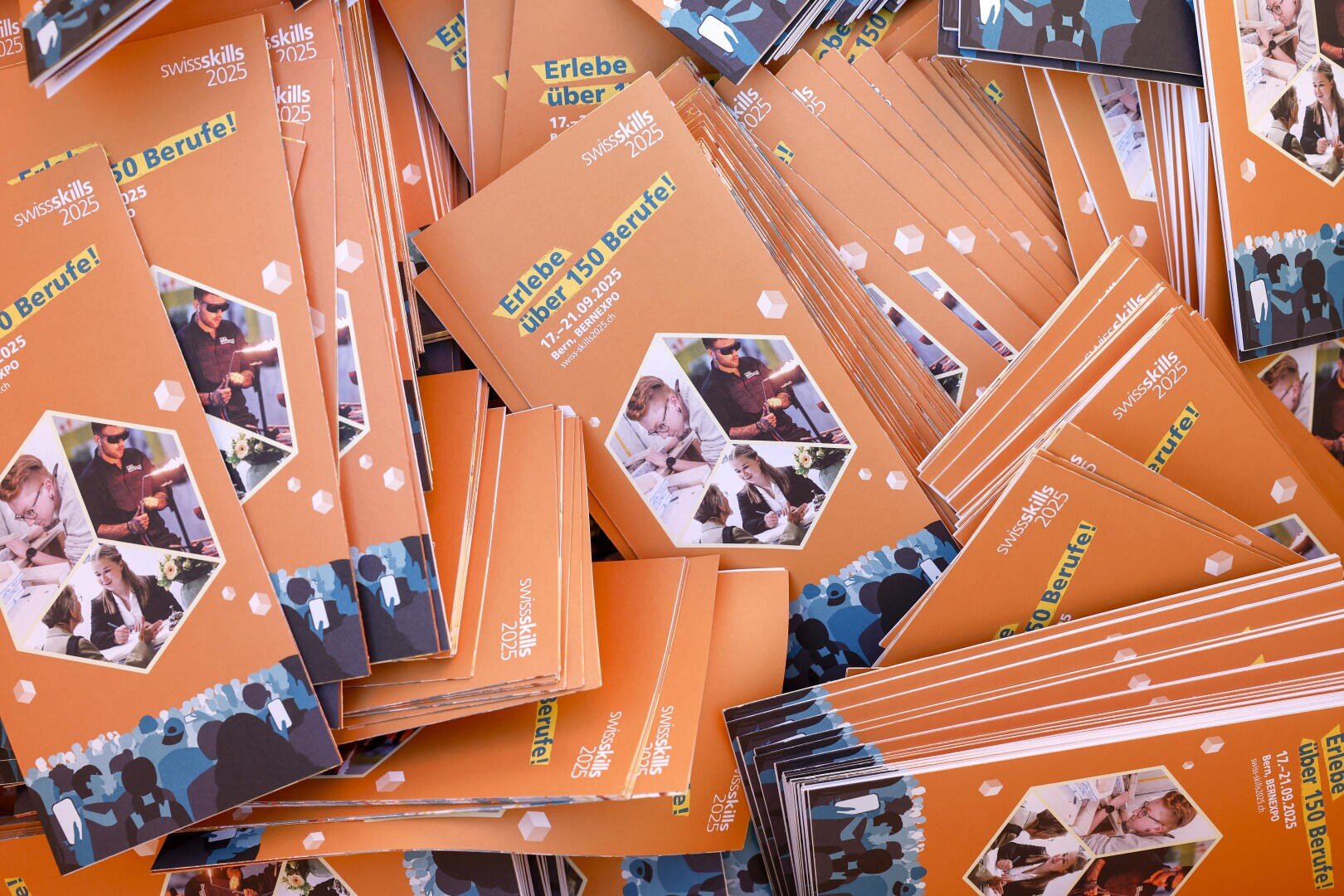 Ein grosser Stapel orangefarbener Broschüren für SwissSkills 2025 mit sechseckigen Fotoausschnitten, blauen Silhouetten und grünem Text, der für die Veranstaltung wirbt, mit der Überschrift Erlebe über 150 Berufe! auf dem Umschlag.