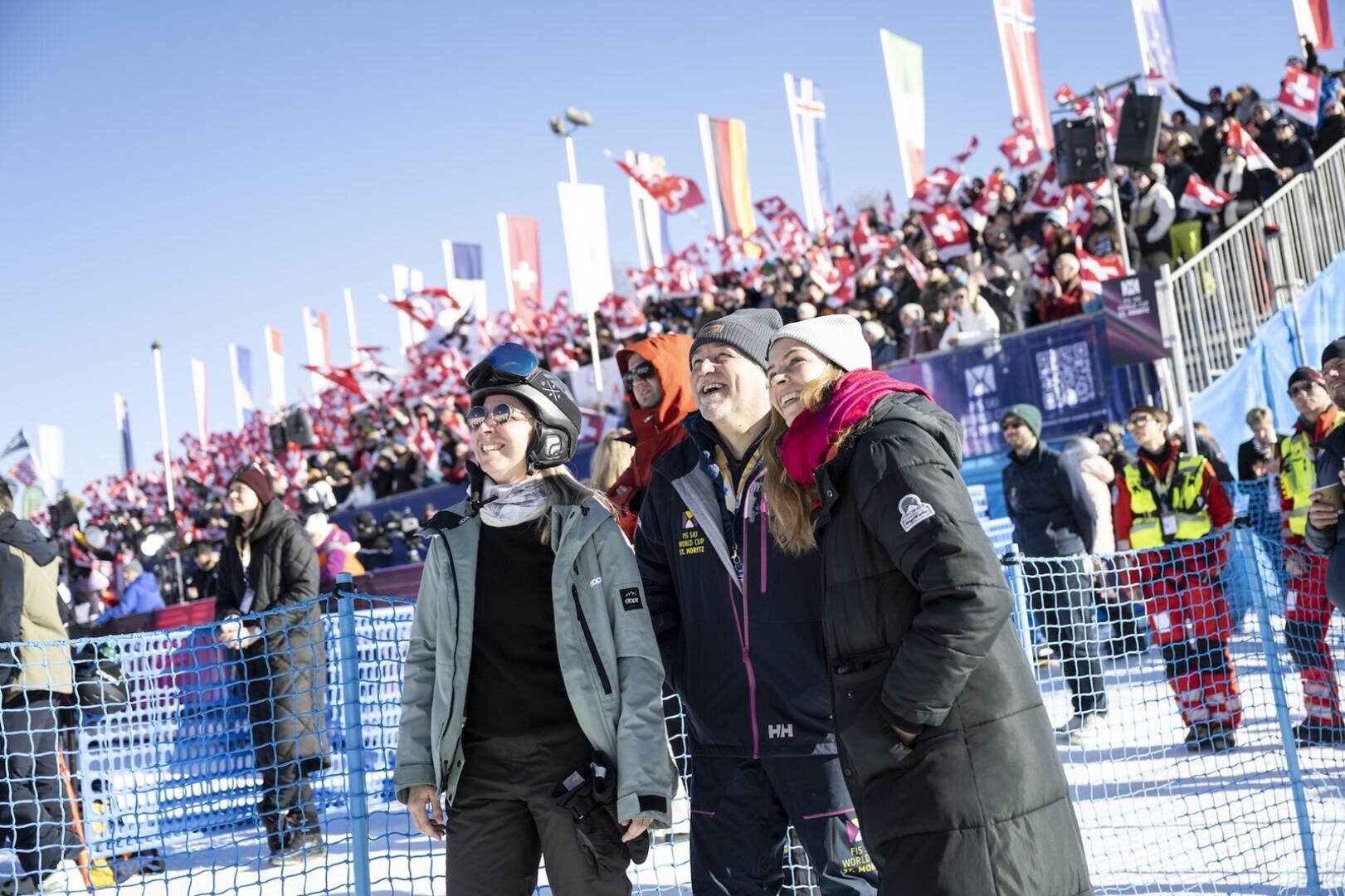 Drei Personen der HF Tourismus & Management stehen lächelnd beim FIS Skiweltcup St. Moritz im Schnee im Freien, hinter ihnen eine große jubelnde Menschenmenge. Bunte Fahnen und Menschen in Winterkleidung füllen die Tribünen.