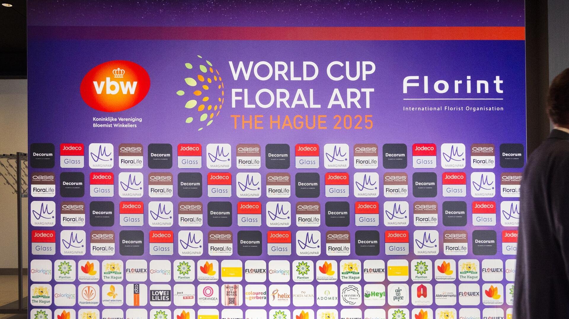 Ein farbenfroher Veranstaltungshintergrund zeigt die Logos des World Cup Floral Art The Hague 2025 mit den Logos der Sponsoren und der Organisationen vbw und Florint in einem lila-orangefarbenen Farbverlaufsdesign.