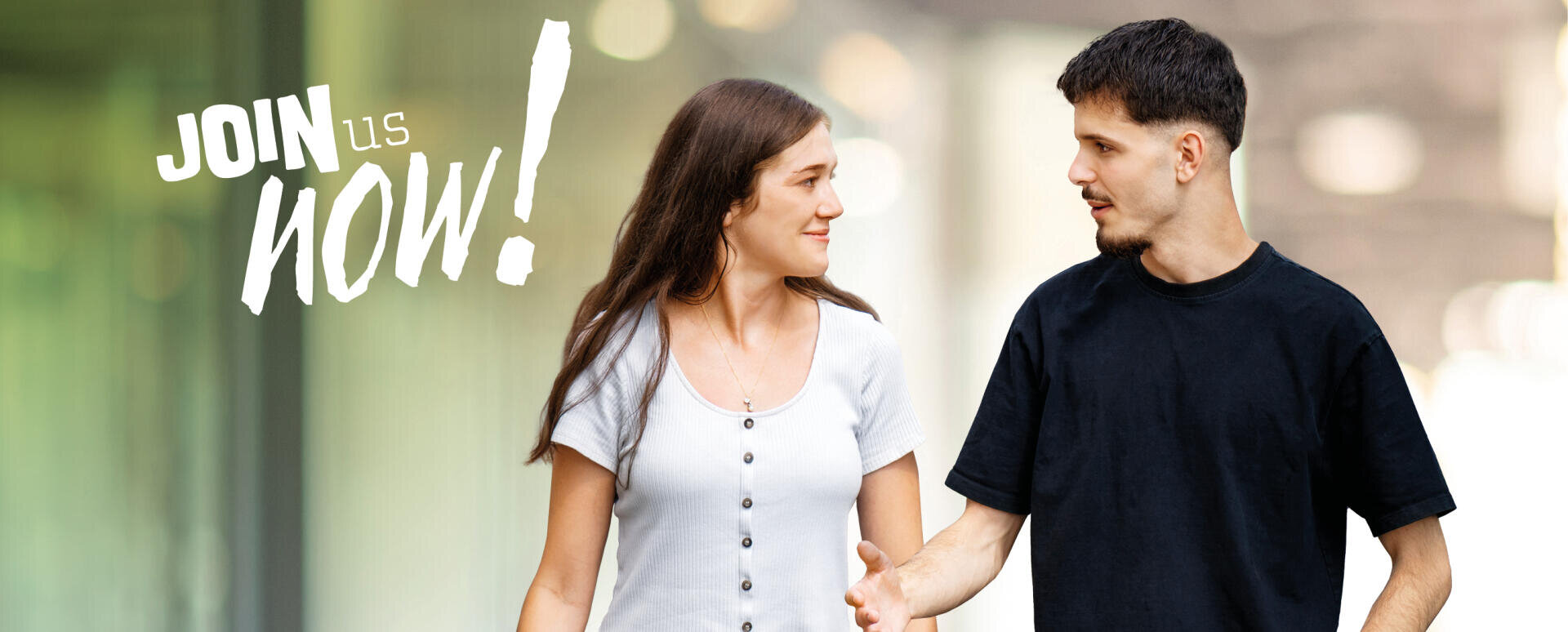 Ein junger Mann und eine junge Frau gehen gemeinsam im Freien spazieren, unterhalten sich und lächeln. Auf der linken Seite steht in fetter weißer Schrift: JOIN us now! Der Hintergrund ist unscharf und hebt die beiden Personen hervor.