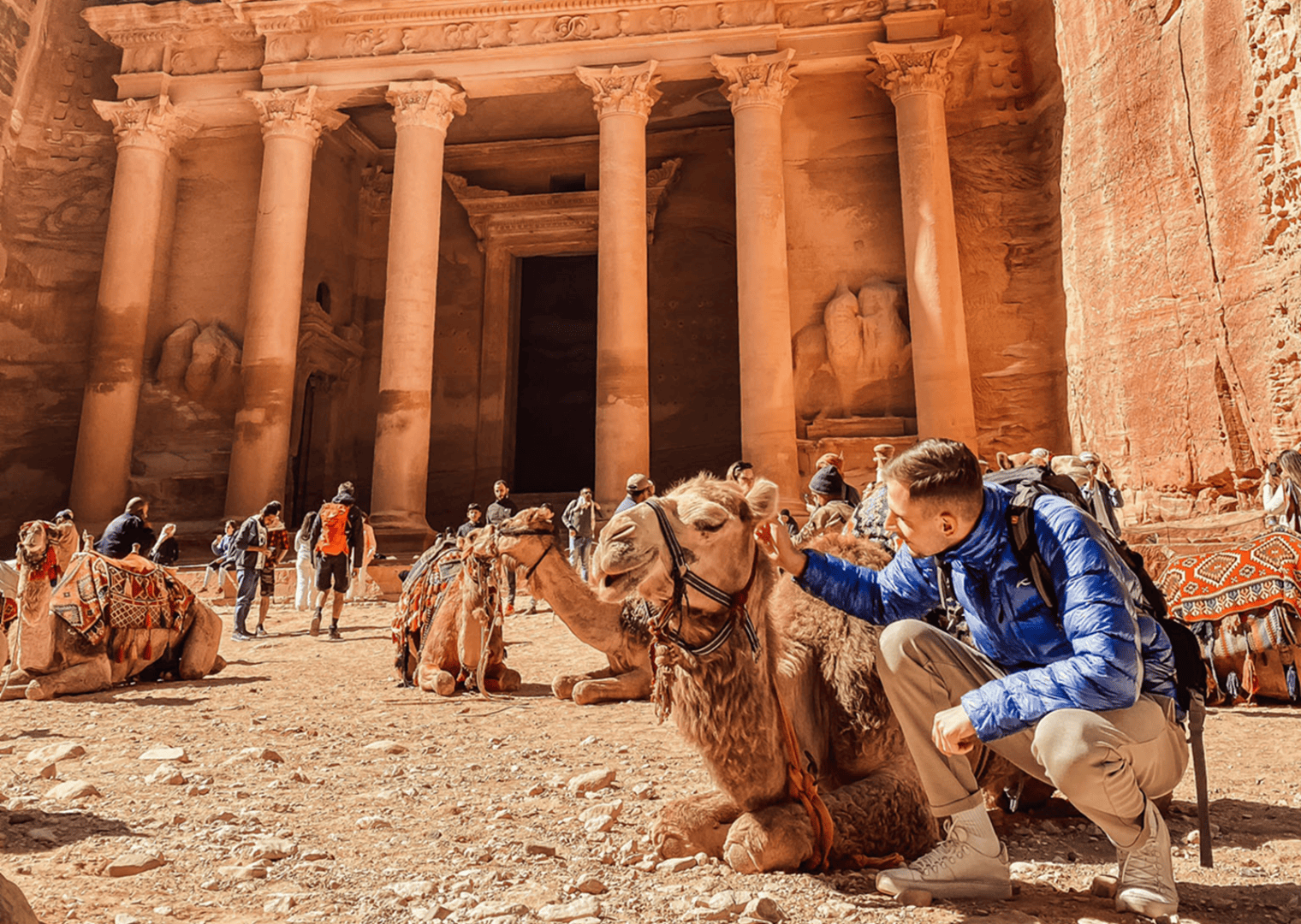 Vor der antiken Steinfassade von Petra in Jordanien sitzt ein Tourismusstudent der HF Tourismus & Mangement (HFT Graubünden) in einer blauen Jacke und streichelt ein Kamel, während in der Nähe weitere Touristen und Kamele zu sehen sind und sonnenbeschienene Felswände die Szene umrahmen.