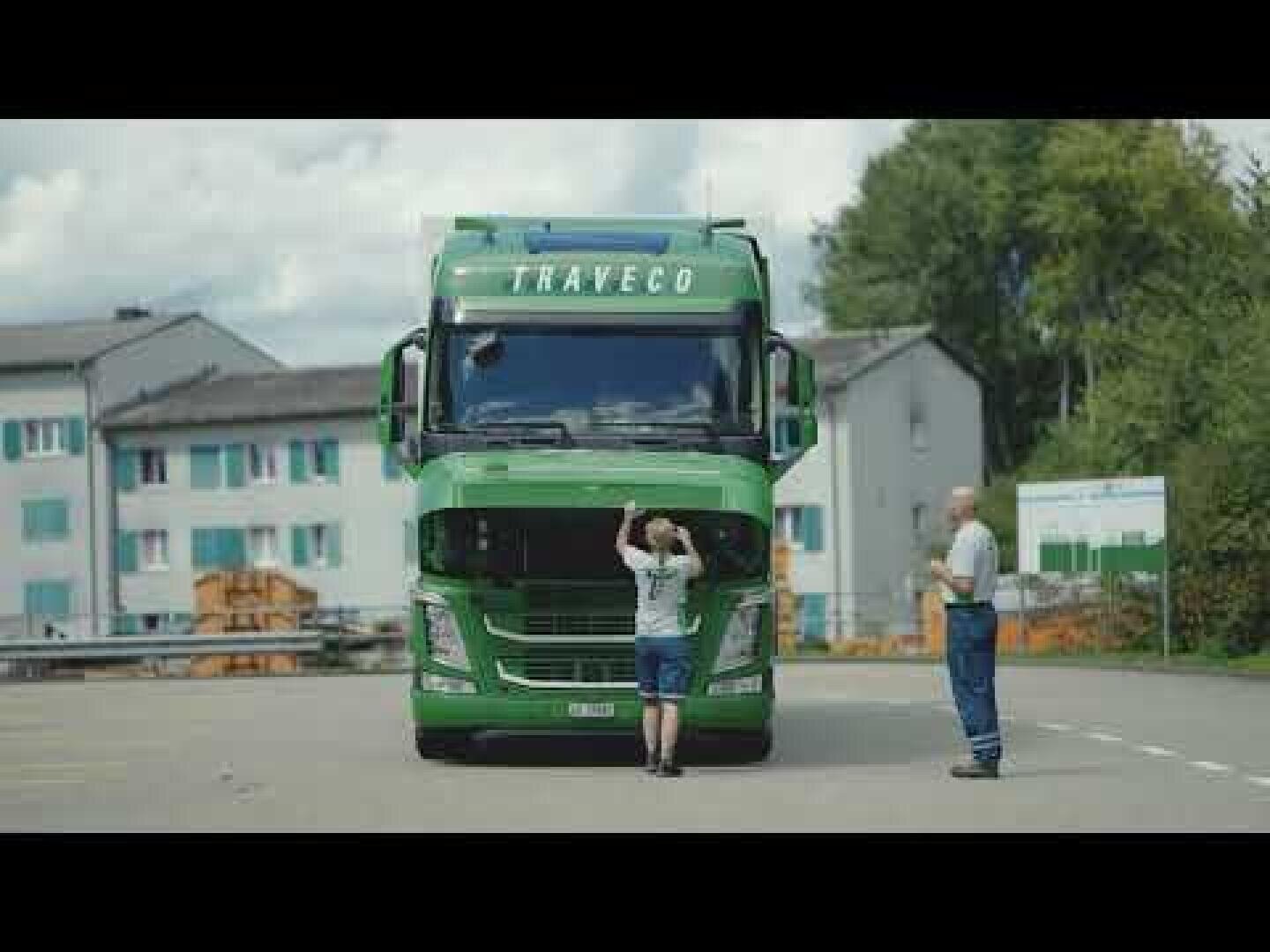 Zwei Personen stehen vor einem großen grünen TRAVECO-Lkw; eine Person schaut unter die angehobene Motorhaube des Lkws, während die andere daneben steht. Im Hintergrund sind Wohnhäuser und Bäume zu sehen.