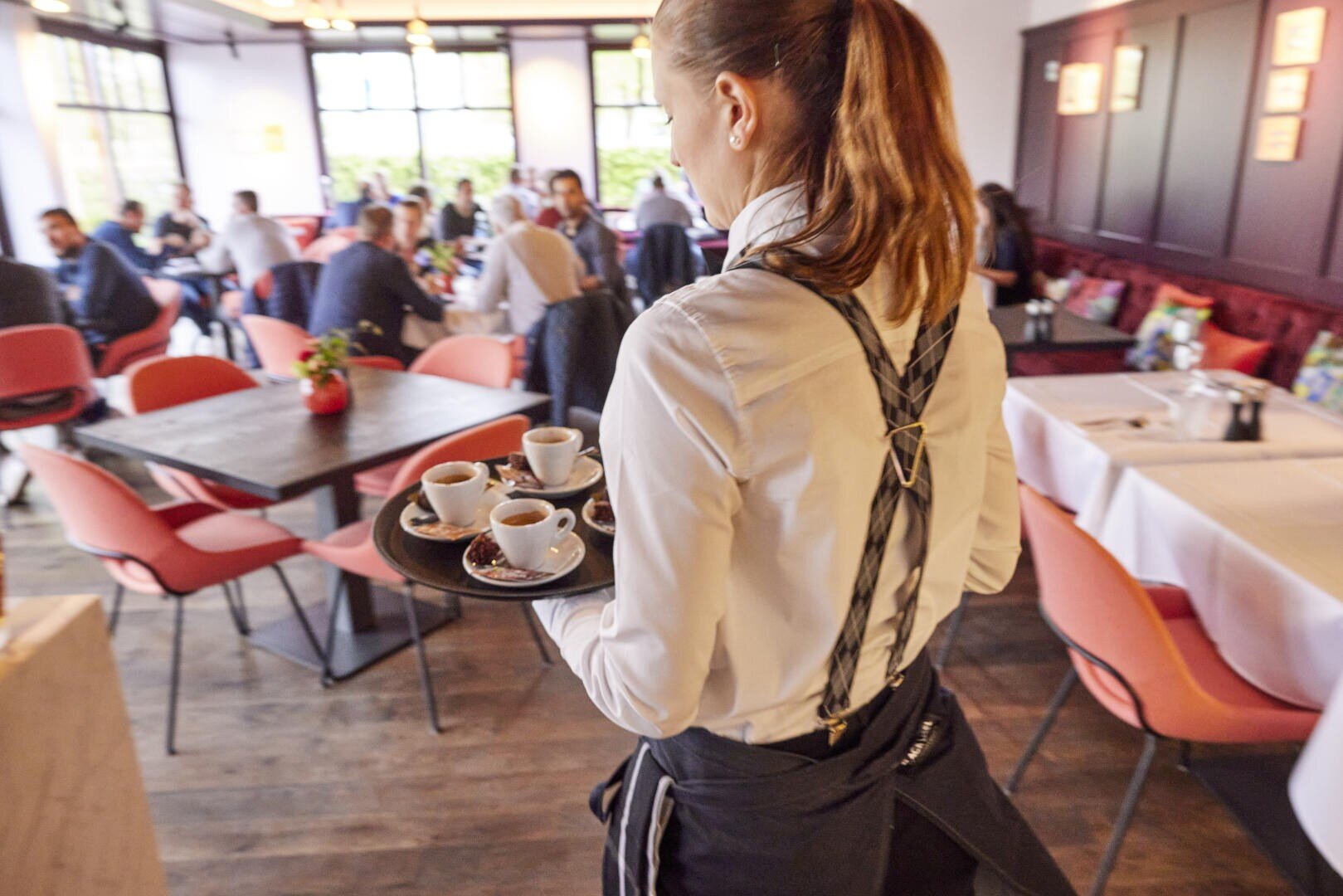 Ein Kellner im weißen Hemd trägt ein Tablett mit Kaffeebechern und kleinen Desserts durch ein lebhaftes, modernes Restaurant mit roten Stühlen und Tischen, an denen Menschen sitzen.