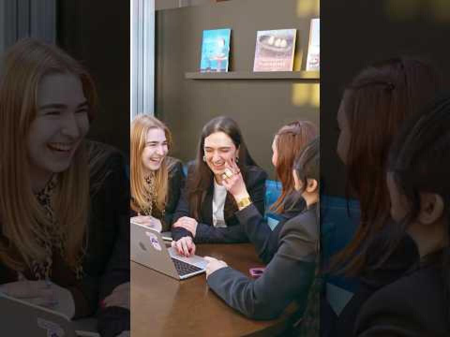 Vier junge Frauen sitzen um einen Tisch herum, lachen und unterhalten sich, während sie auf einen Laptop schauen. Sie scheinen sich in einem Café oder einem Lernraum zu befinden; im Hintergrund sind Bücher in einem Regal zu sehen.