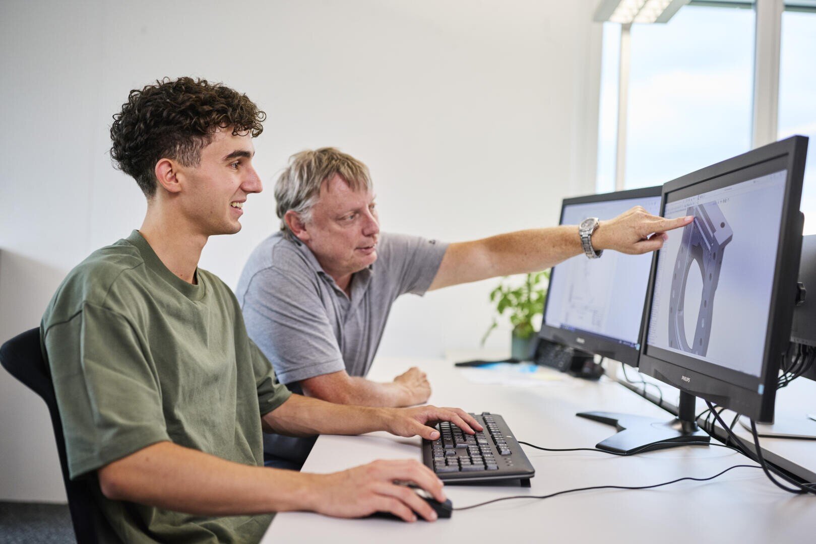 Zwei Männer sitzen an einem Schreibtisch mit Computermonitoren. Der jüngere Mann tippt, während der ältere Mann auf eine technische Zeichnung auf dem Bildschirm zeigt, was auf eine Anleitung oder Instruktion in einer modernen Büroumgebung hindeutet.