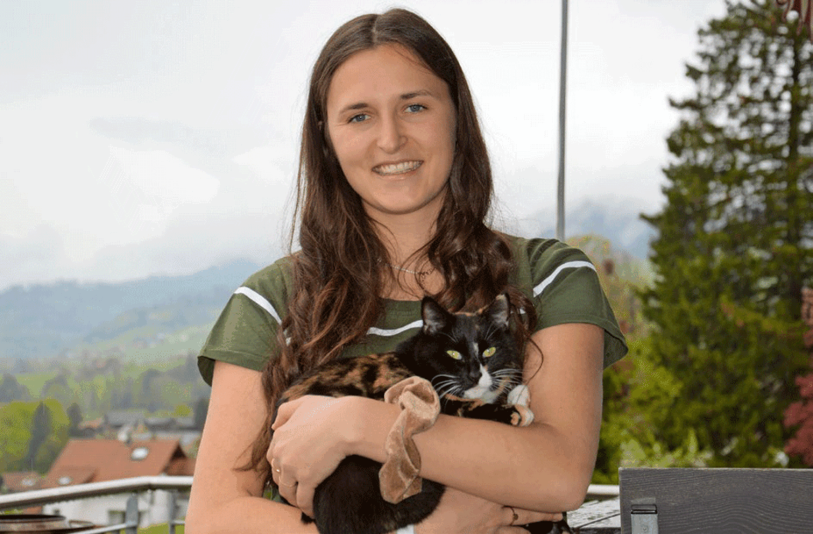 Eine Frau mit langen braunen Haaren lächelt, während sie eine schwarz-orangefarbene Katze auf dem Arm hält, vor dem Hintergrund von Bäumen, Hügeln und Häusern.