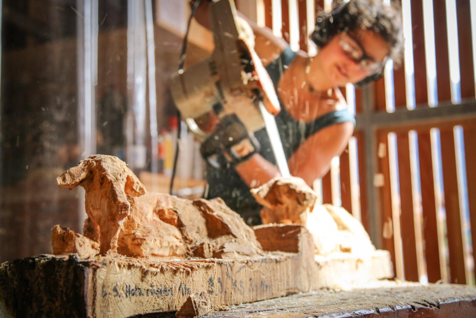 Eine Person in Schutzkleidung schnitzt mit einer Kettensäge eine Holzskulptur von Tieren, während in der sonnendurchfluteten Werkstatt Holzspäne um sie herumfliegen.