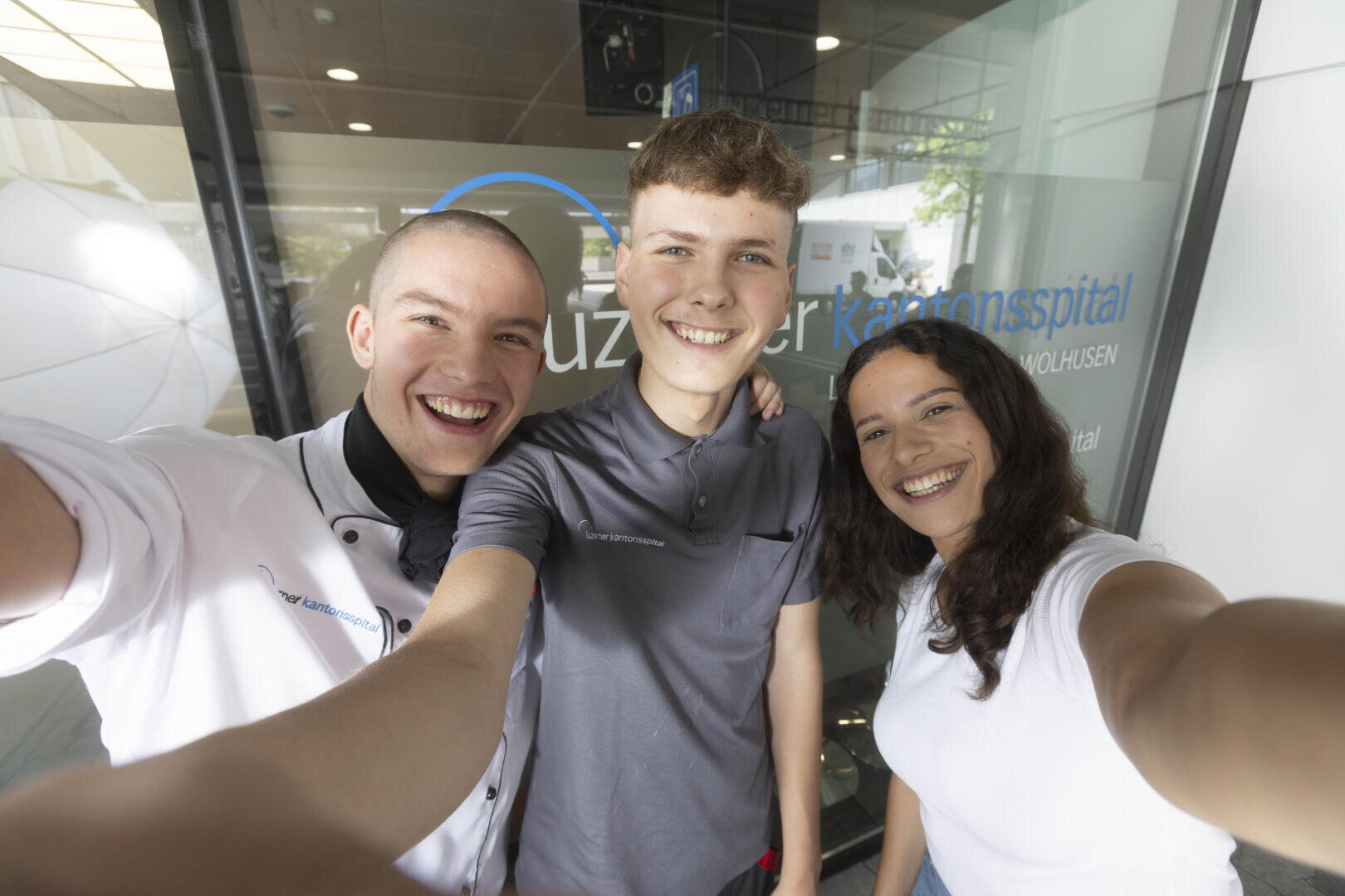 Drei junge Erwachsene lächeln und machen gemeinsam ein Selfie vor einer Glastür mit der Aufschrift "Luzerner Kantonsspital". Sie wirken glücklich und tragen legere Arbeitsuniformen.