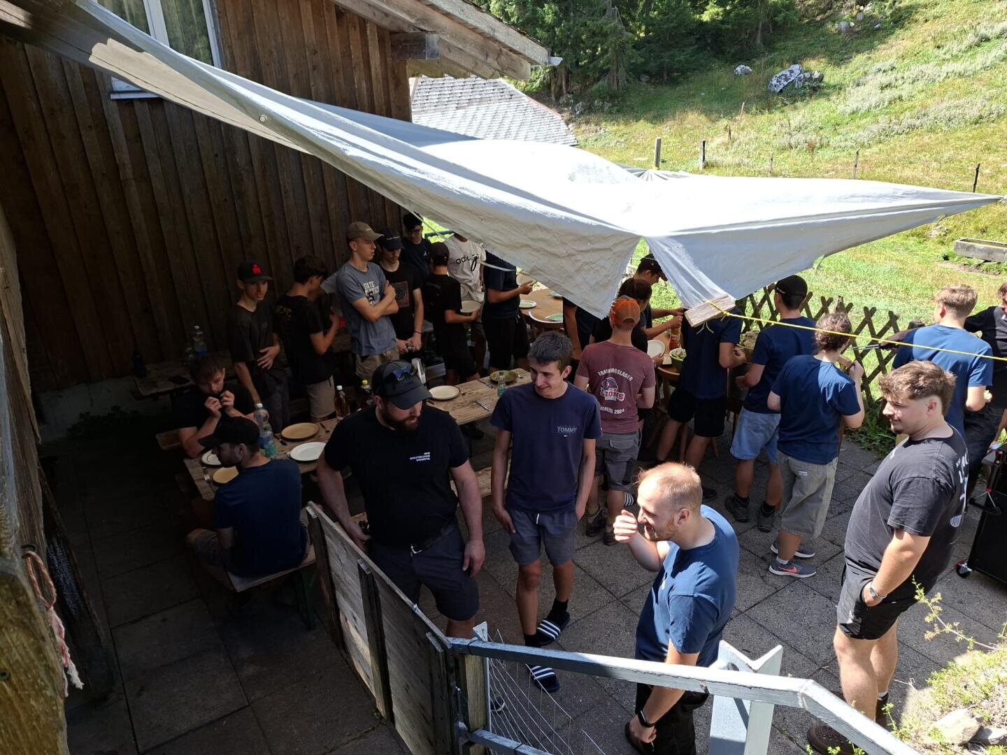 Eine Gruppe von Menschen hat sich unter einer weißen Plane neben einem Holzgebäude versammelt und isst und unterhält sich an einem sonnigen Tag im Freien. Einige sitzen mit Tellern an Tischen, während andere stehen und sich unterhalten.