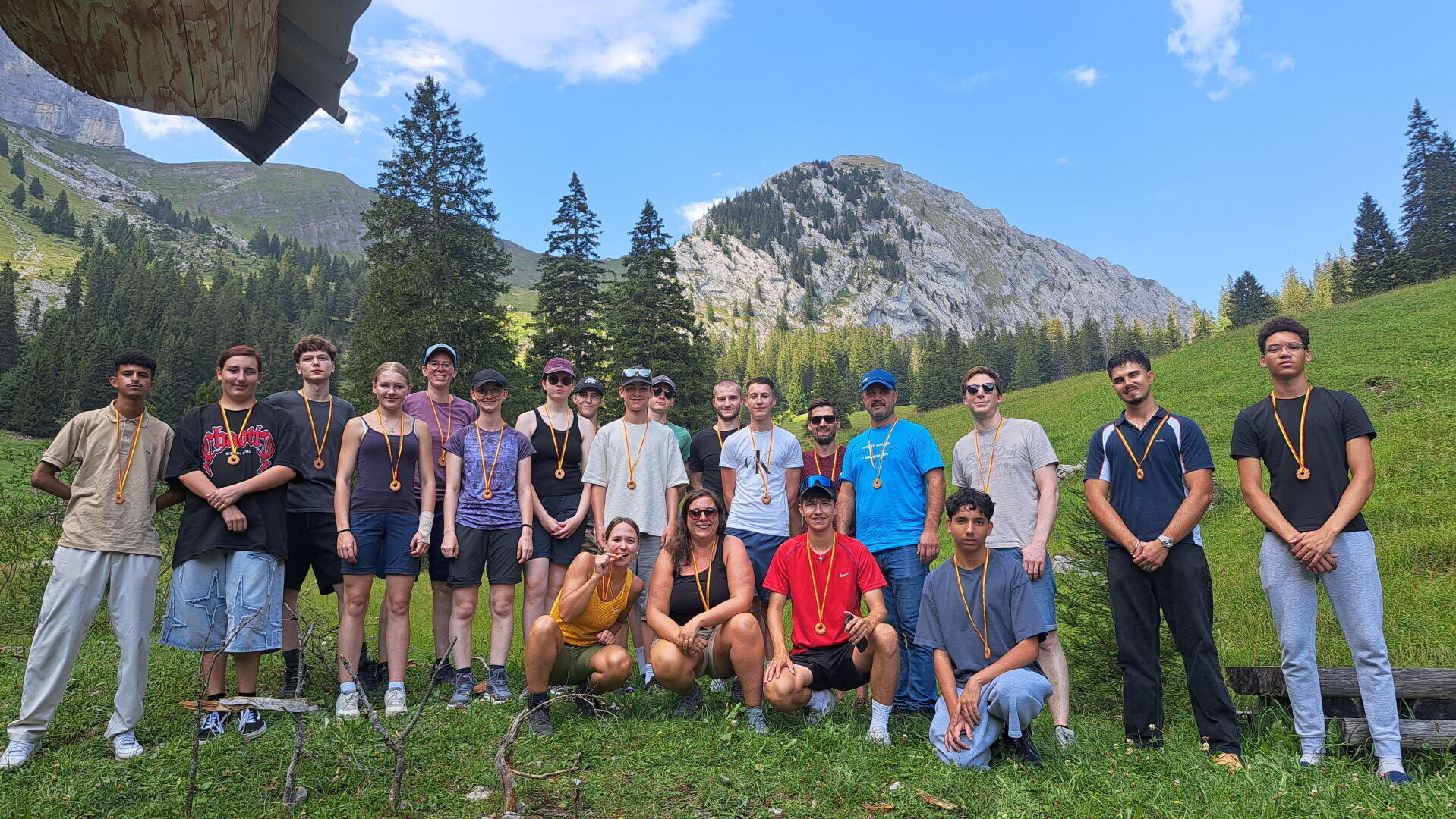 Eine Gruppe junger Erwachsener und Jugendlicher, von denen einige Medaillen tragen, posiert im Freien in einer bergigen, grasbewachsenen Landschaft mit Bäumen und blauem Himmel im Hintergrund.
