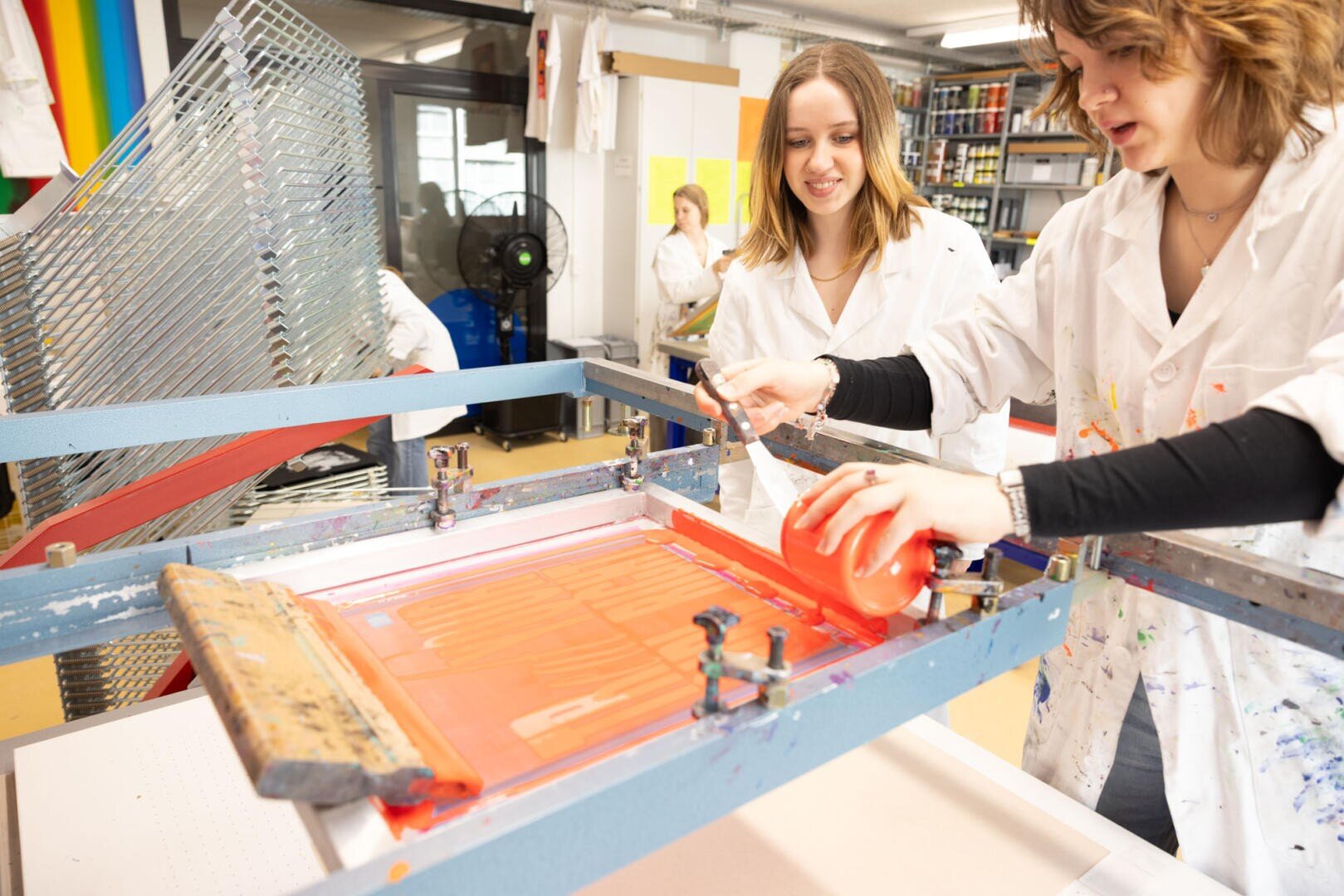 Zwei Frauen in weißen Laborkitteln arbeiten gemeinsam an einem Siebdruckprojekt. In einem gut beleuchteten Kunststudio gießen sie lächelnd leuchtend orangefarbene Farbe auf ein Sieb.