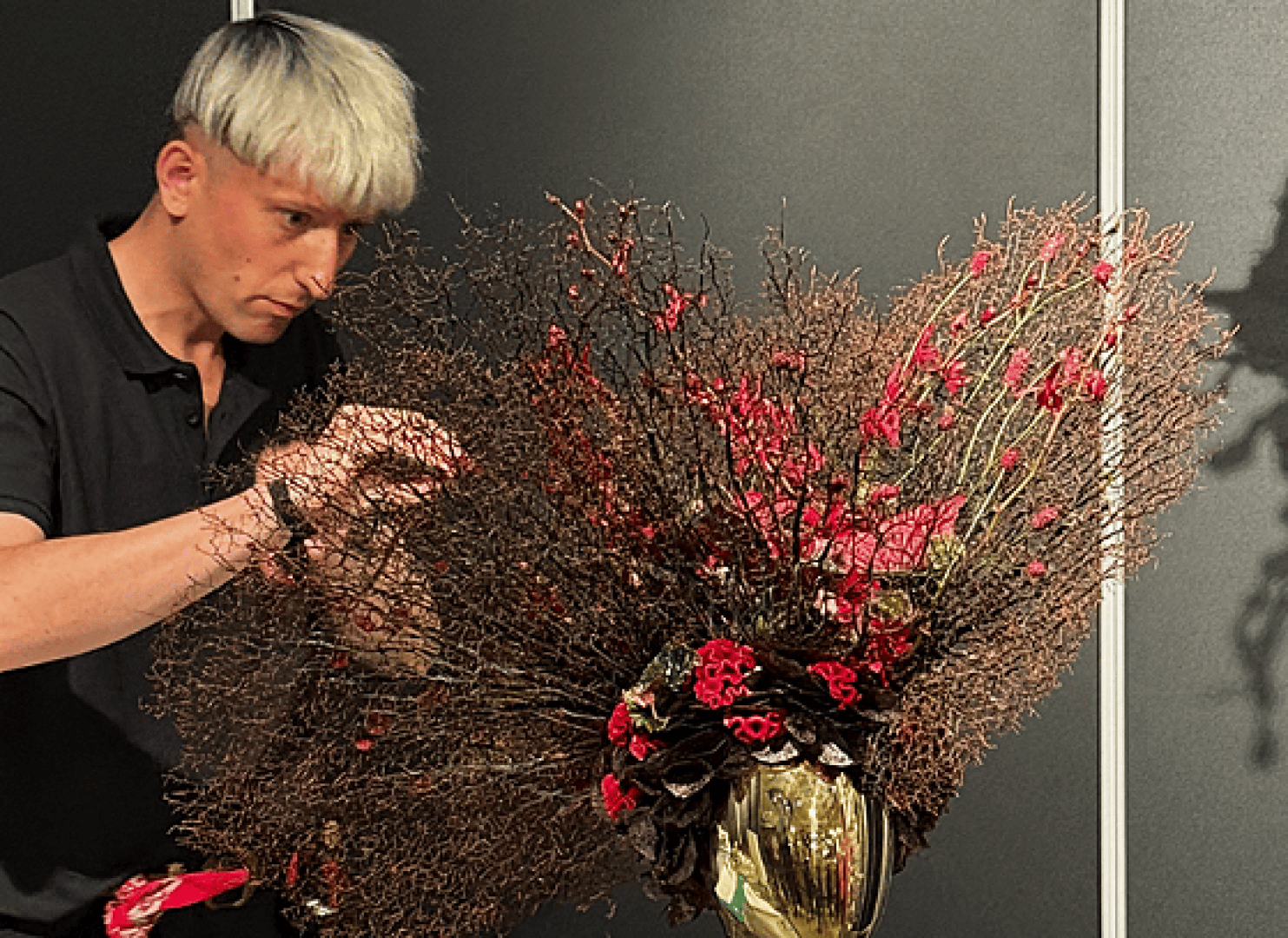 Eine Person mit kurzem, silbernem Haar arrangiert einen kunstvollen Blumenschmuck mit roten Blumen und dichten, braunen Zweigen in einer goldenen Vase vor einem dunklen Hintergrund.