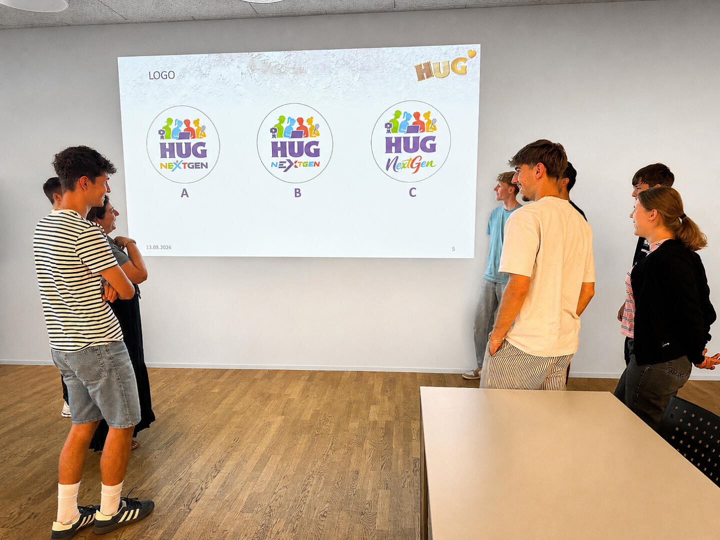 Sieben Personen stehen in einem Raum und betrachten einen Bildschirm, auf dem drei farbenfrohe Logo-Entwürfe mit den Bezeichnungen A, B und C für „HUG NextGen“ zu sehen sind.