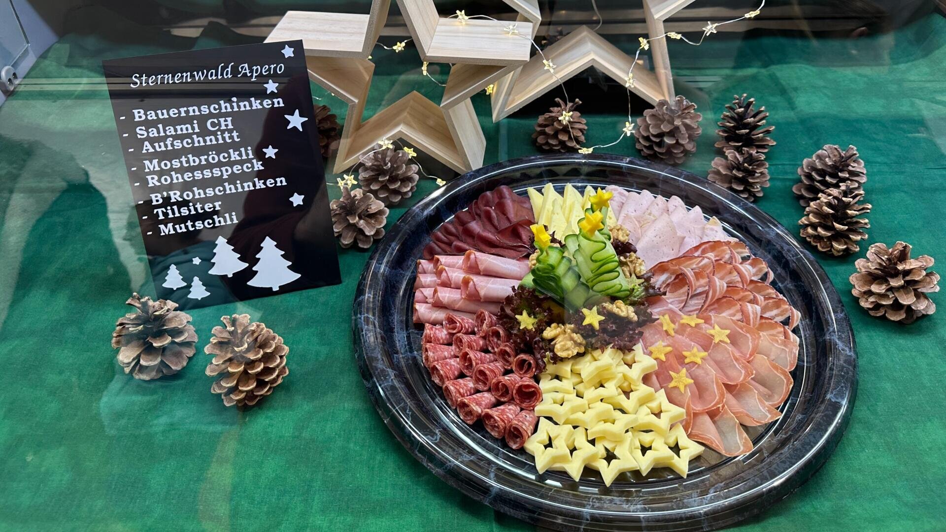 Auf einem grünen Tuch ist eine Platte mit verschiedenen Fleisch- und Käsescheiben angerichtet, umgeben von Tannenzapfen und Holzsternen. Auf einem Schild sind die Produkte aufgeführt: Bauernschinken, Salami, Aufschnitt, Mostbröckli, Rohessspeck, Rohschinken, Tilsiter, Mutschli.
