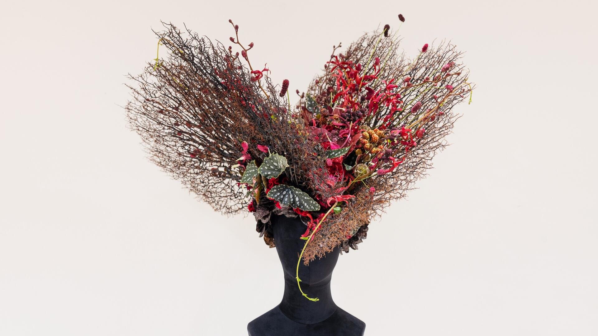 Eine schwarze Schaufensterpuppe trägt einen kunstvollen Kopfschmuck aus getrockneten Zweigen, roten Blüten und grünen Blättern, die sich weit vor einem schlichten hellen Hintergrund ausbreiten.