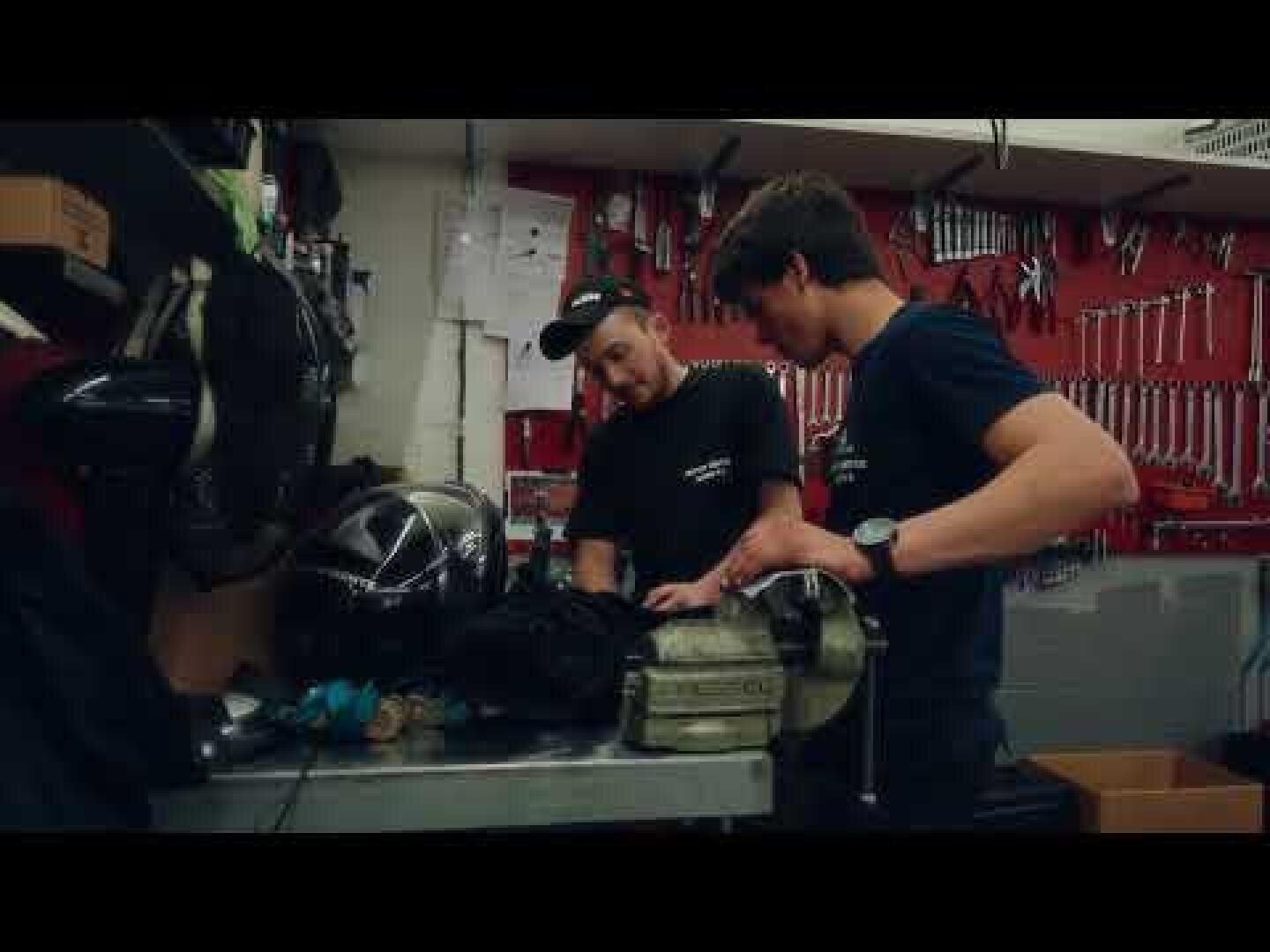 Zwei junge Männer arbeiten in einer Werkstatt gemeinsam an einem mechanischen Teil an einer unübersichtlichen Werkbank, umgeben von Werkzeugen, die an einer roten Wand im Hintergrund hängen.