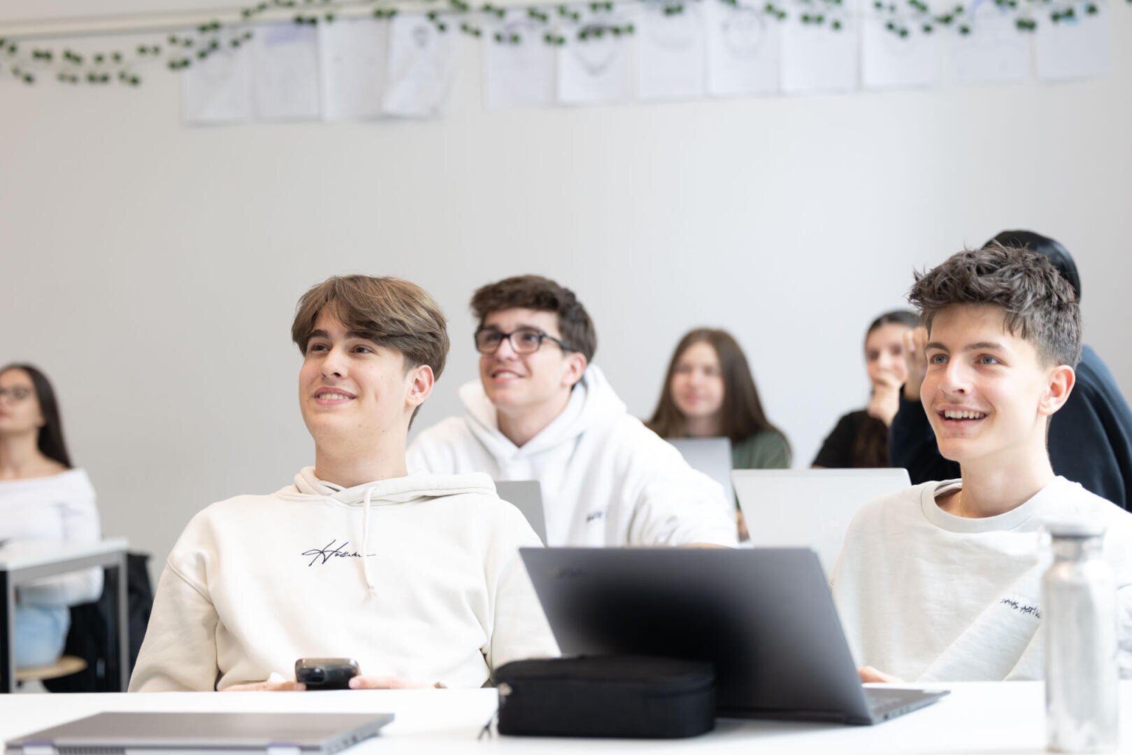 Eine Gruppe von Schülern sitzt in einem hellen Klassenzimmer, lächelt und blickt nach vorne. Zwei Jungen im Vordergrund haben Laptops vor sich geöffnet. Andere Schüler sind im Hintergrund zu sehen.